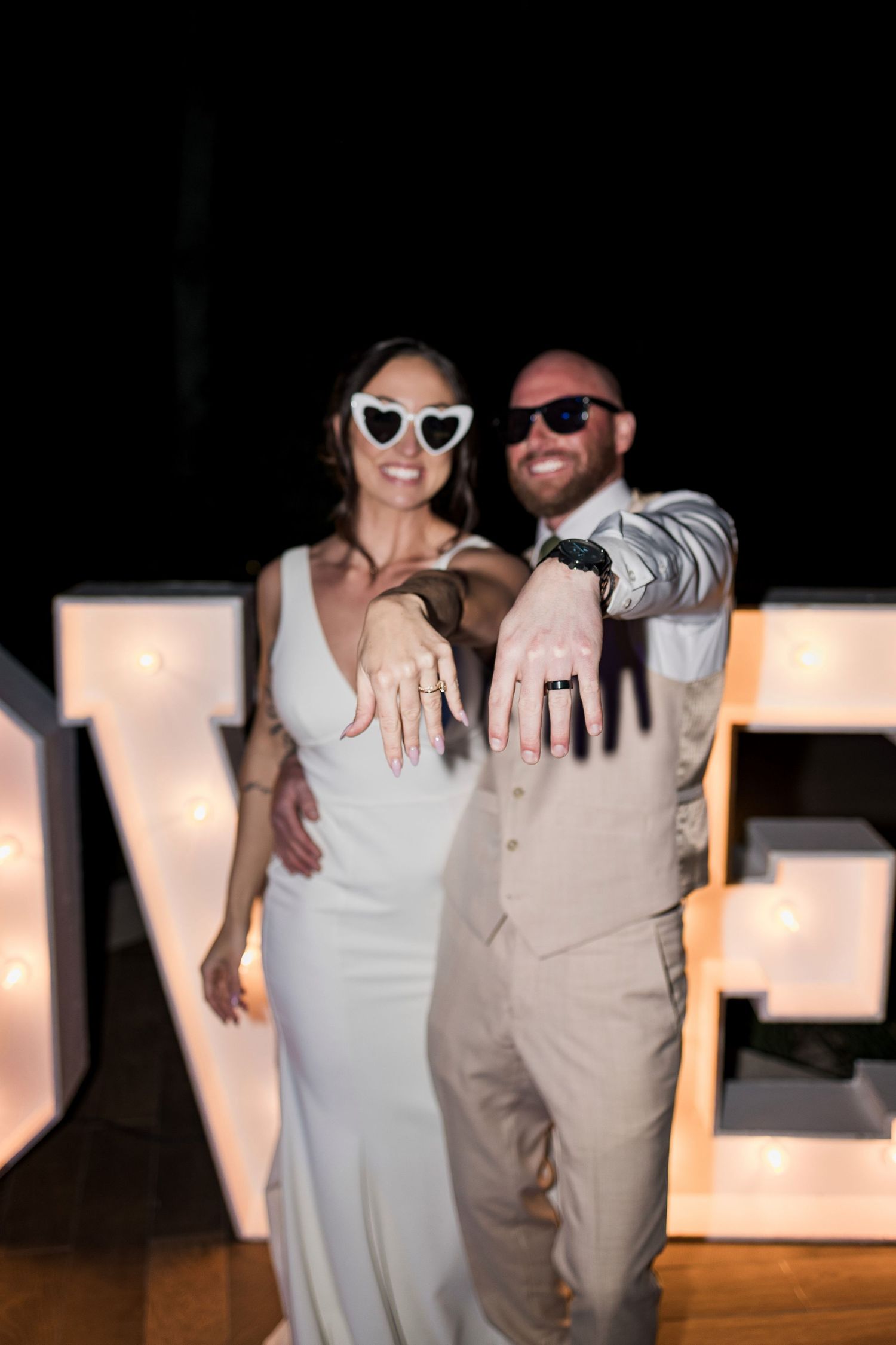 Newlyweds in sunglasses showing off wedding rings in front of illuminated LOVE letters at their reception.