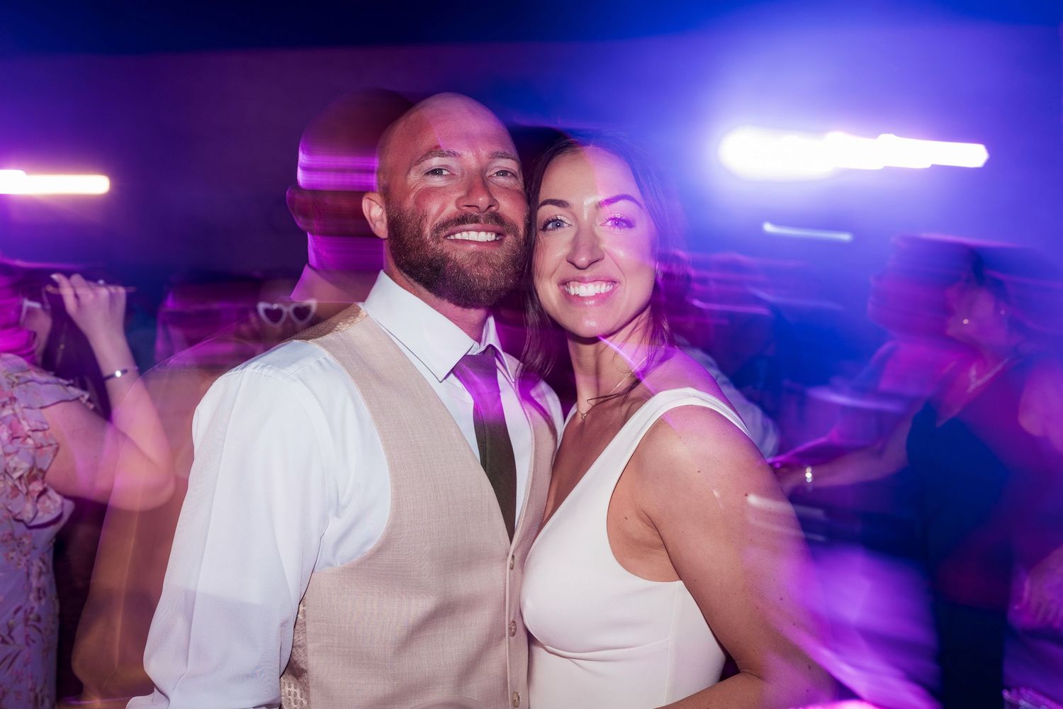Smiling couple poses together on a colorful dance floor with purple lights at a wedding reception.