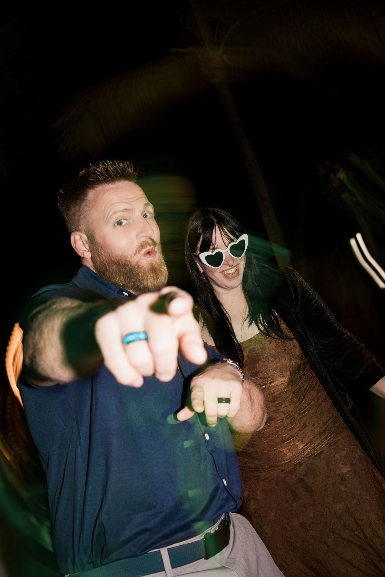 Bearded man in blue shirt pointing at camera with woman in heart sunglasses and green hair behind him at night event.