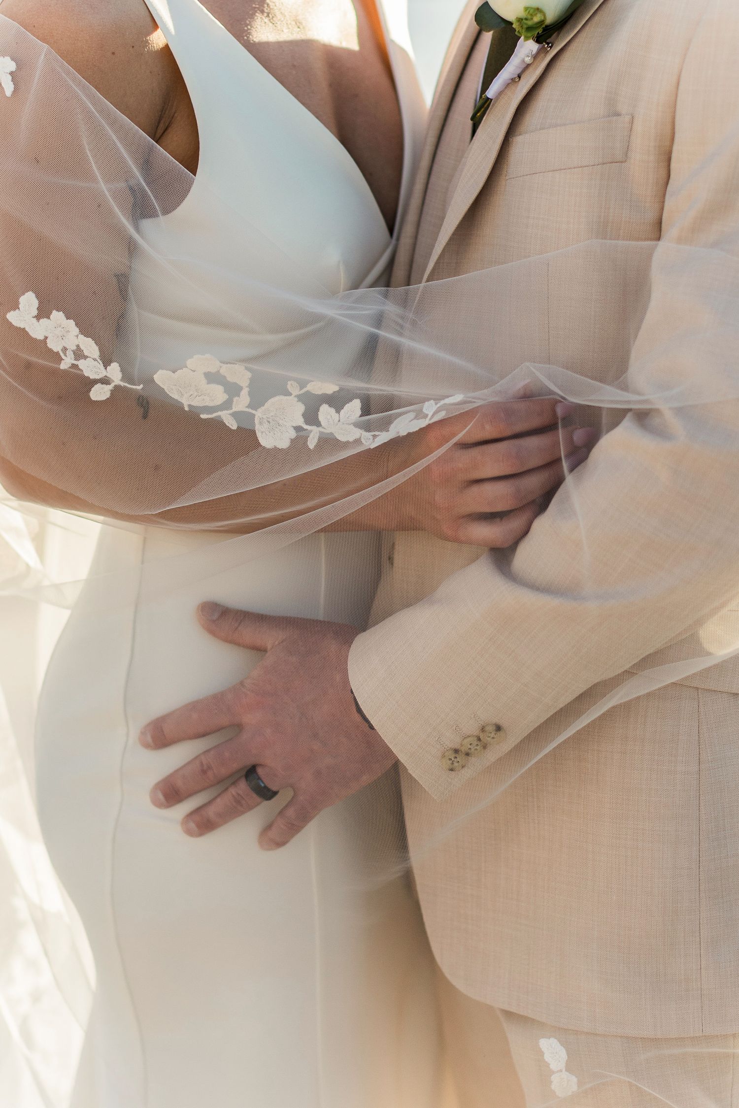 Close-up of bride and groom embracing, showcasing wedding rings, lace veil, and groom's beige suit with floral boutonniere.