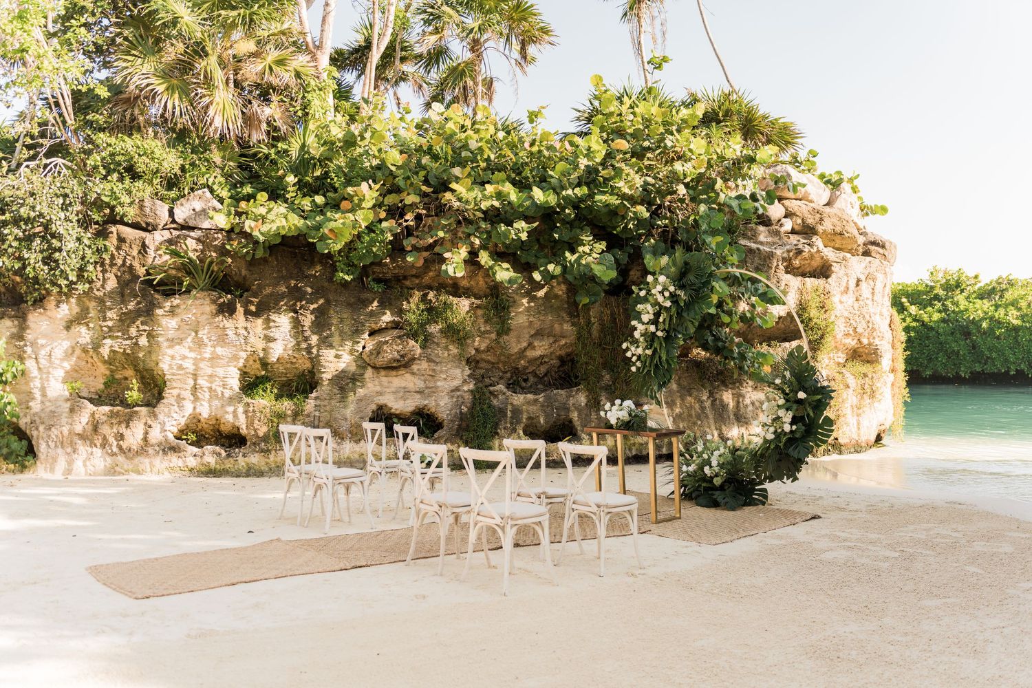 Xcaret Arte Wedding - Small beach wedding setup with white chairs on sand, framed by a lush, vine-covered rocky cliff and turquoise water.