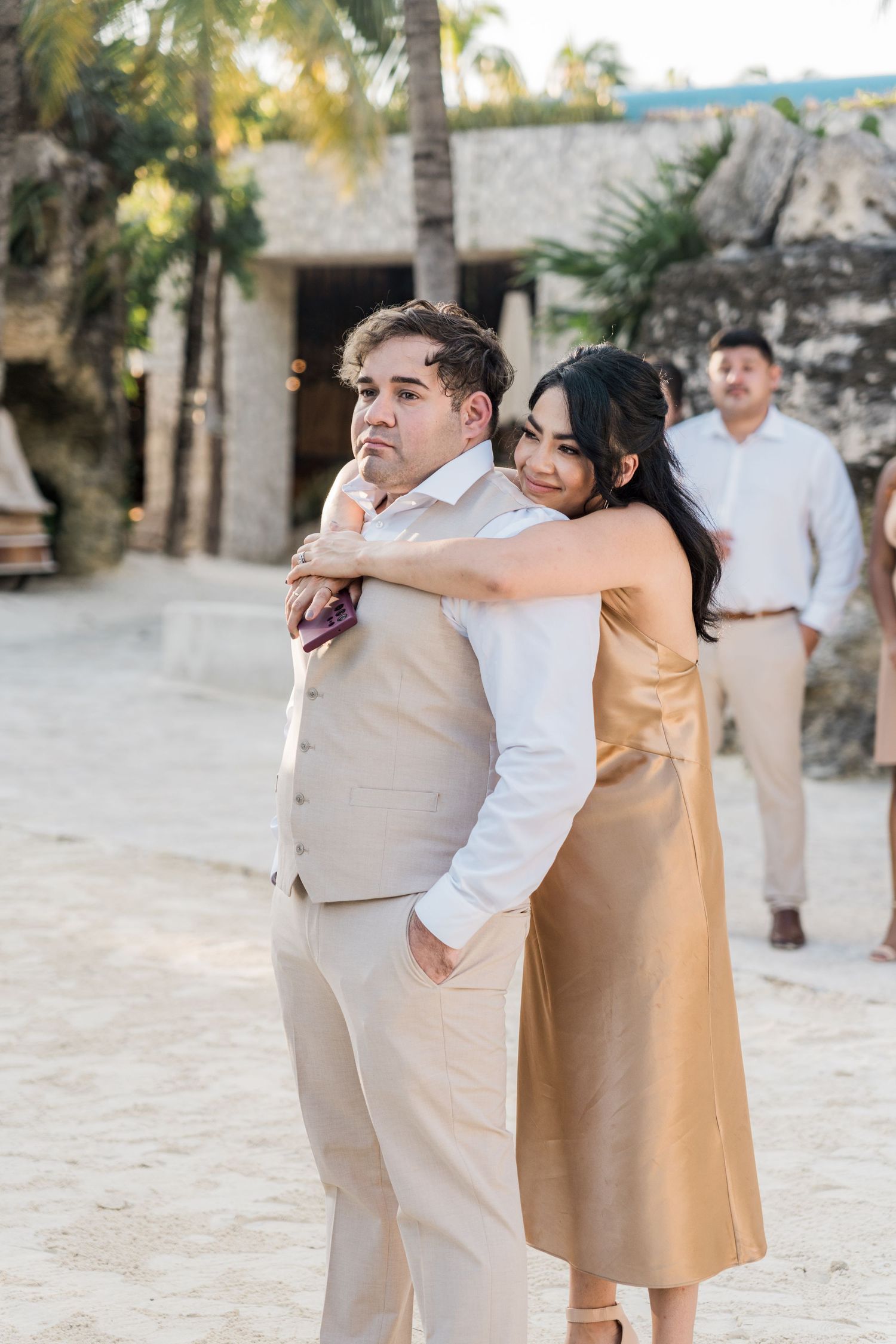 Xcaret Arte Wedding Couple embracing on sandy beach at outdoor wedding venue with palm trees, woman in gold dress hugging man in beige suit.