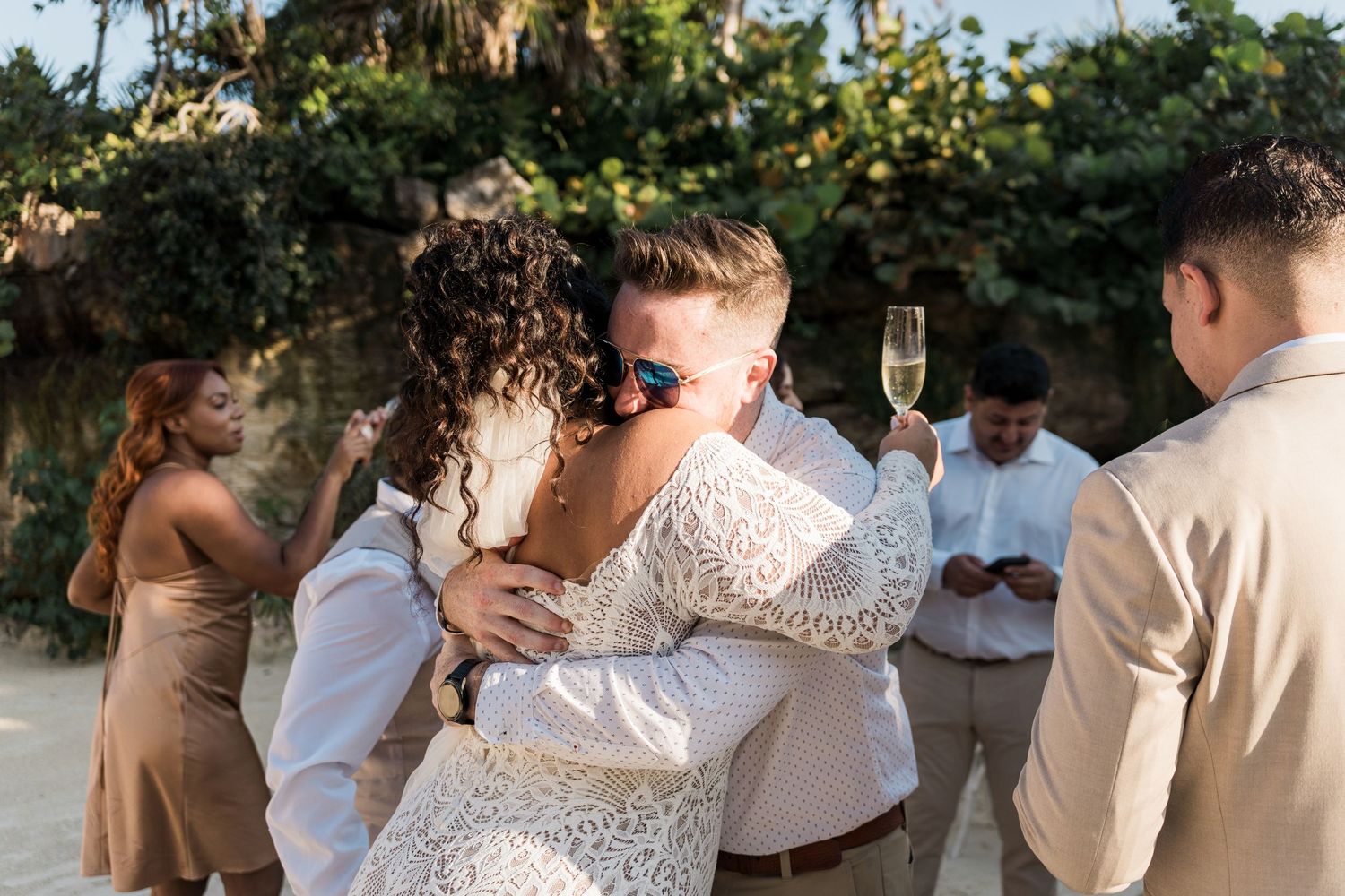 Xcaret Arte Wedding - Bride and groom share a heartfelt hug during outdoor beach wedding ceremony with guests holding champagne.