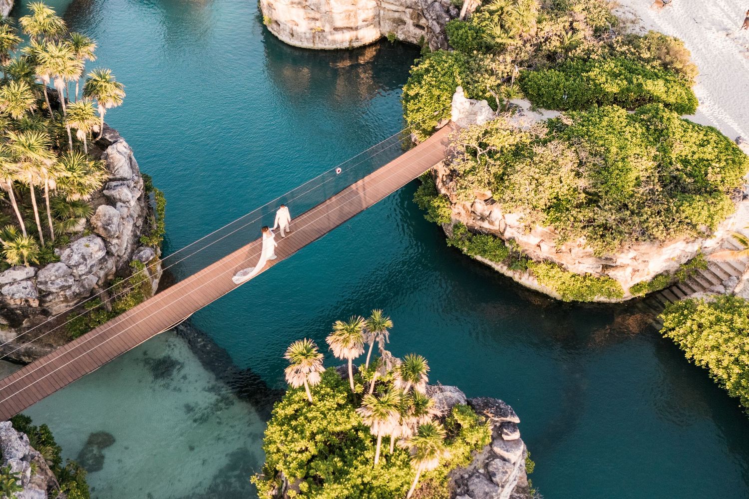 Xcaret Arte Wedding - Aerial view of a suspension bridge over turquoise waters connecting rocky islands with lush tropical palm trees.