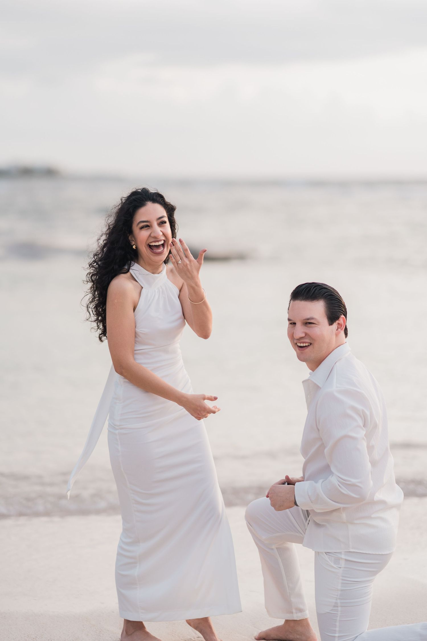 Joyful couple in white outfits on beach, man on one knee proposing as woman shows off engagement ring.