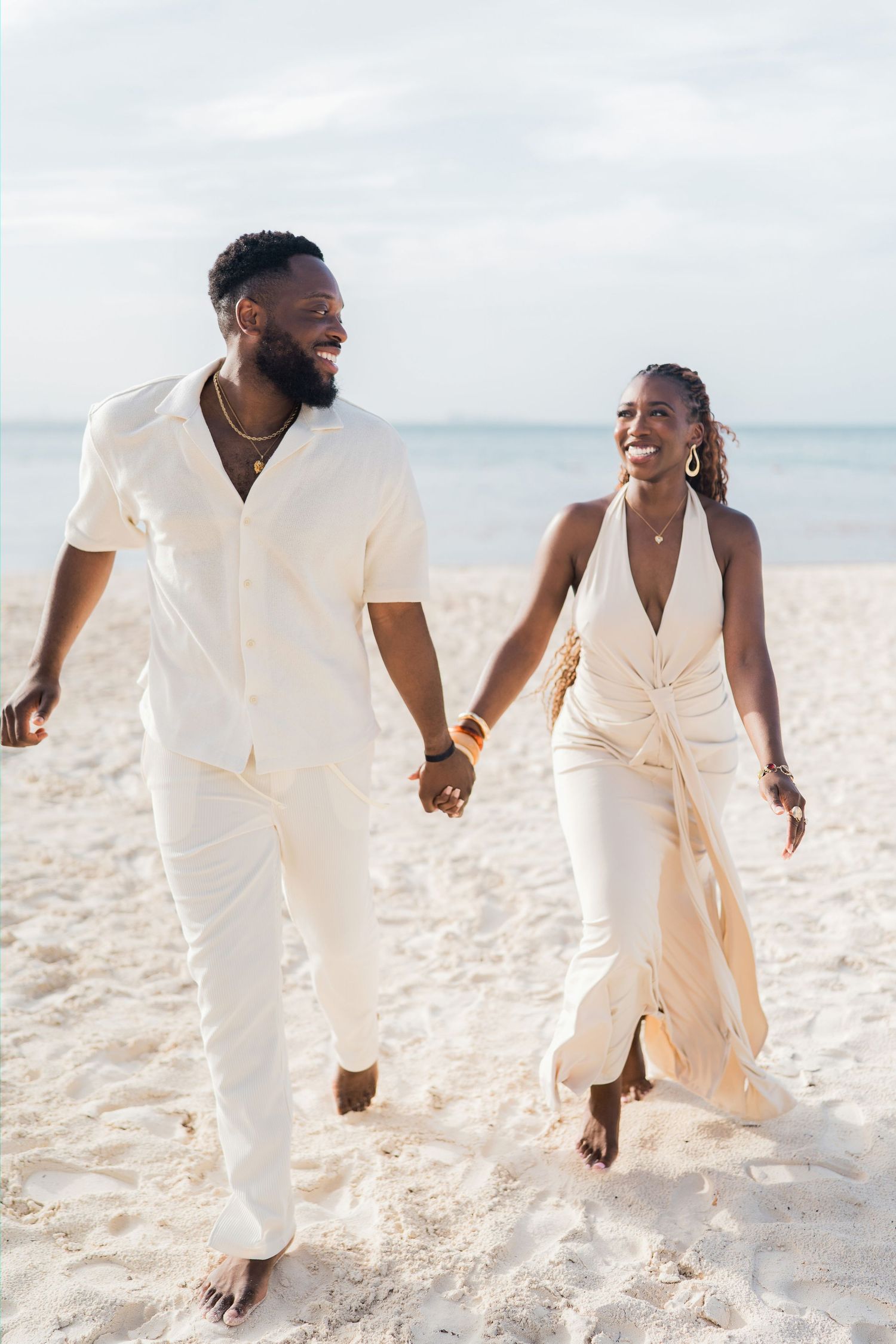 Happy couple in matching cream outfits walking hand-in-hand on a sandy beach with calm ocean waters behind them.