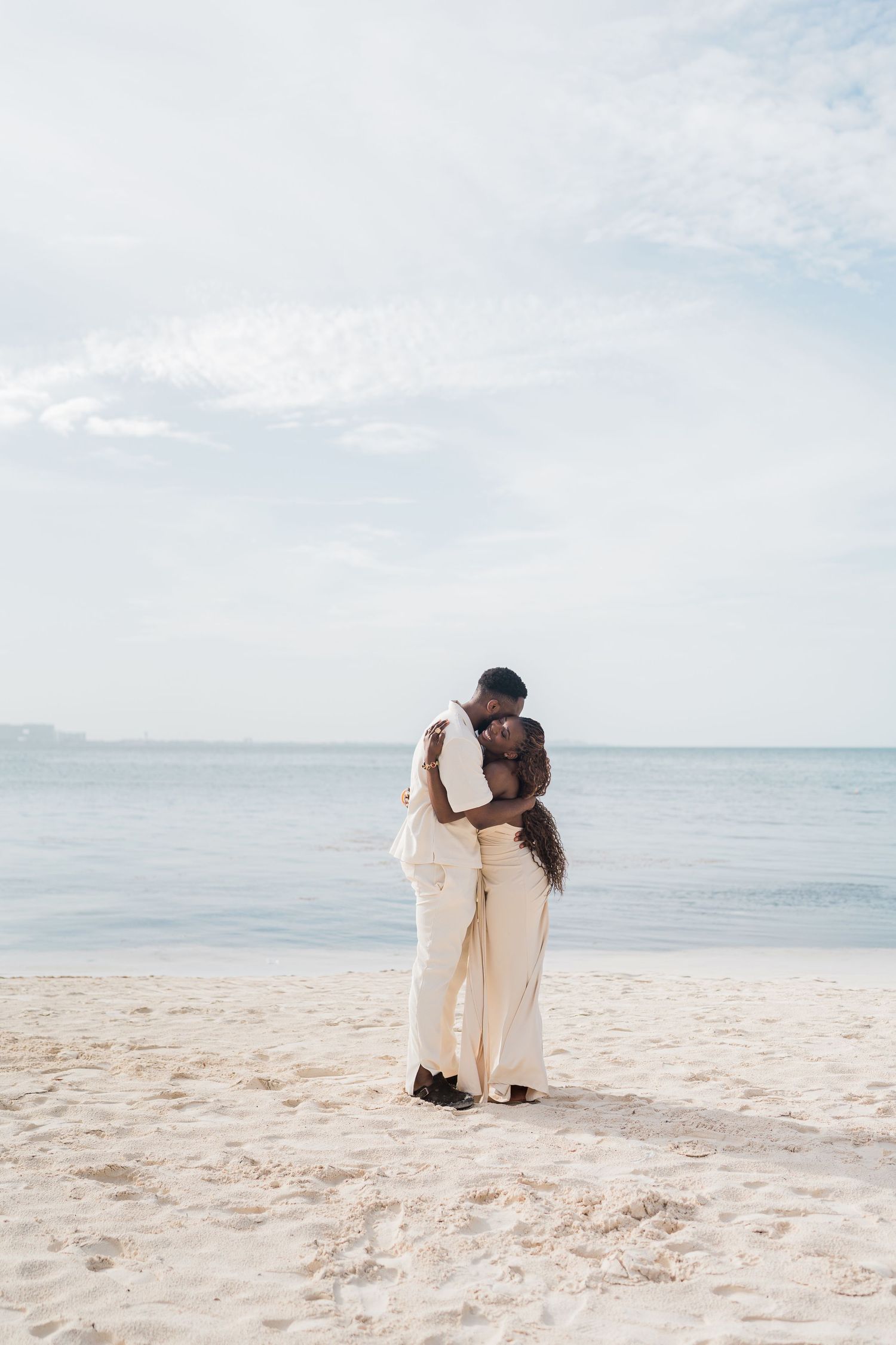 A couple embracing on a white sand beach in matching cream outfits, with calm blue ocean waters stretching behind them.
