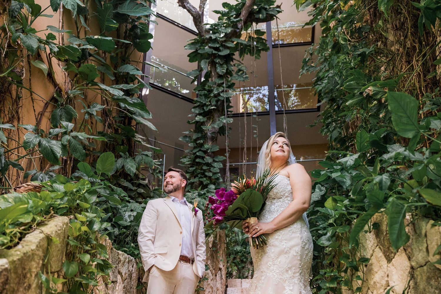 Hotel Xcaret México Wedding - Bride and groom stand among lush greenery indoors, bride holding vibrant purple bouquet