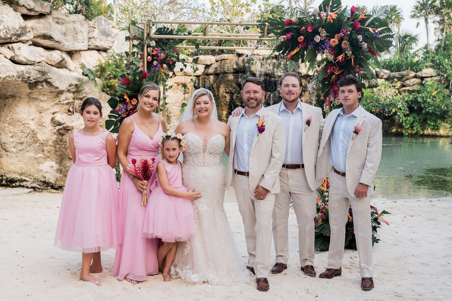 Wedding party poses outdoors near tropical waterfall, bride in white gown with bridesmaids in pink dresses and groomsmen in tan suits.