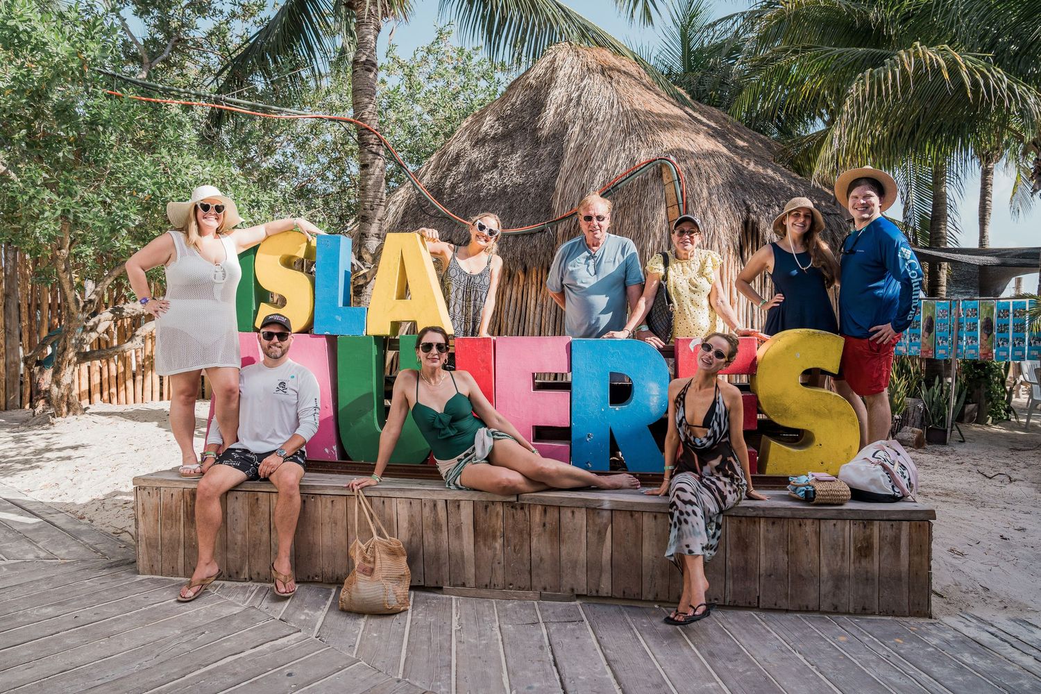 Riviera Maya Multi Day wedding - group posing with colorful Isla Mujeres sign letters on a wooden deck near a thatched hut beach resort.