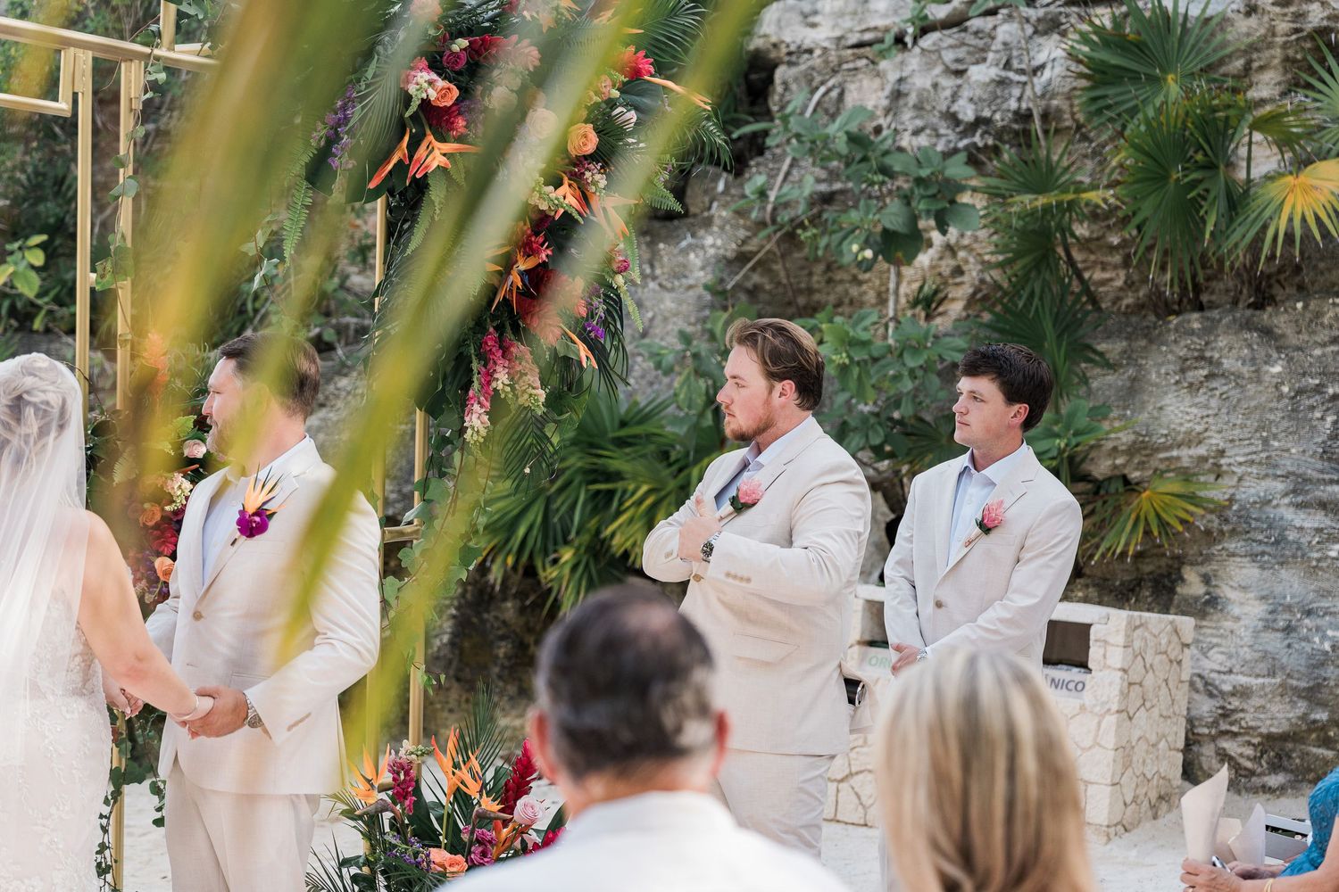 Groomsmen in tan suits stand at a tropical outdoor wedding ceremony with lush greenery and colorful floral arrangements.