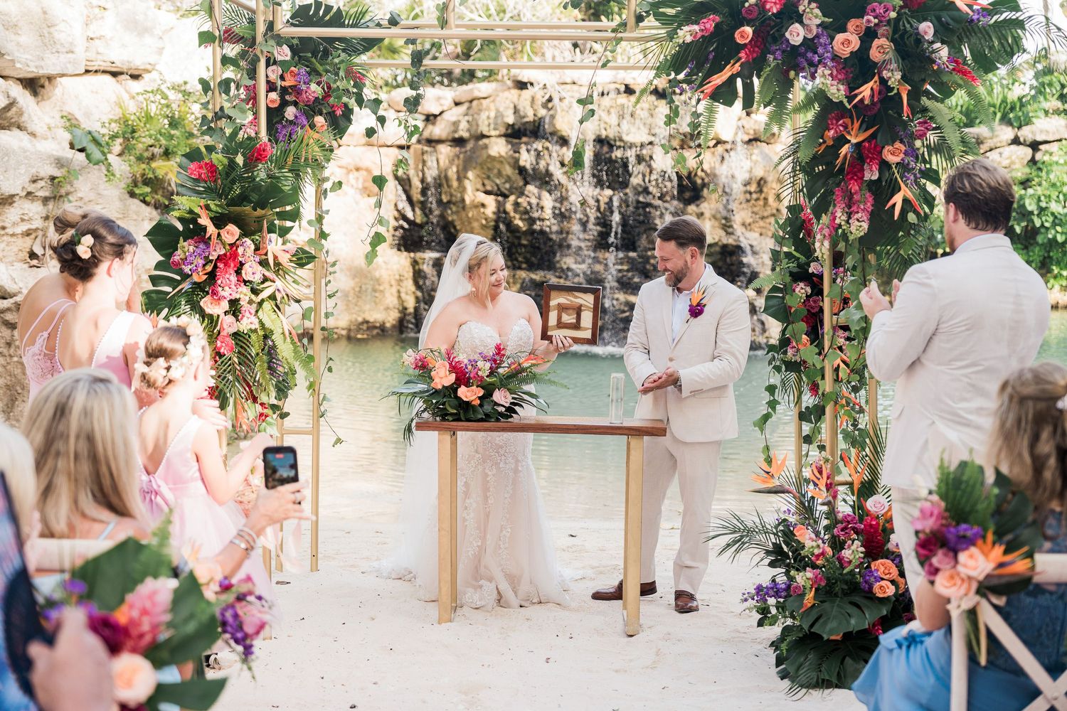 Couple exchanges vows at tropical outdoor wedding ceremony with colorful floral arch and waterfall backdrop.
