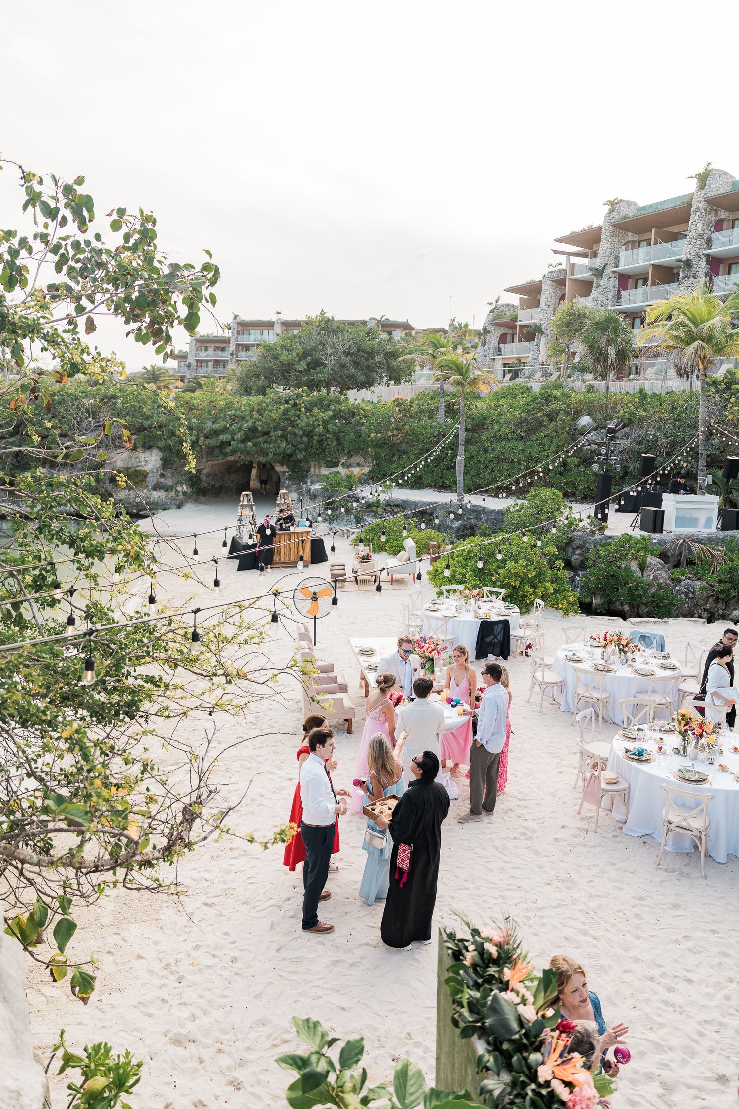 Hotel Xcaret México elegant beach wedding reception with guests mingling on white sand, decorated tables, and string lights near a resort.