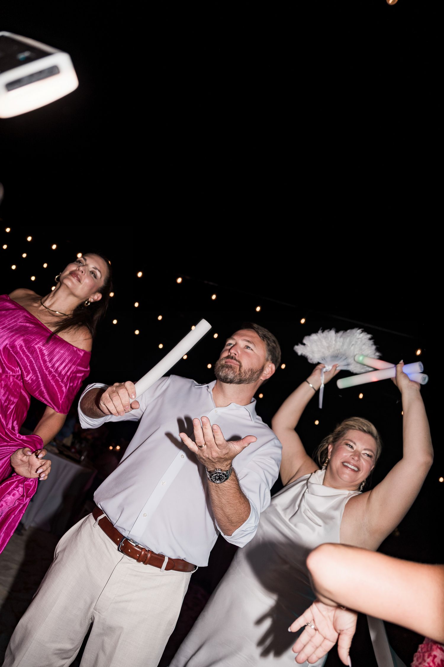 Guests dancing and celebrating at an outdoor evening wedding reception holding glow sticks under string lights.