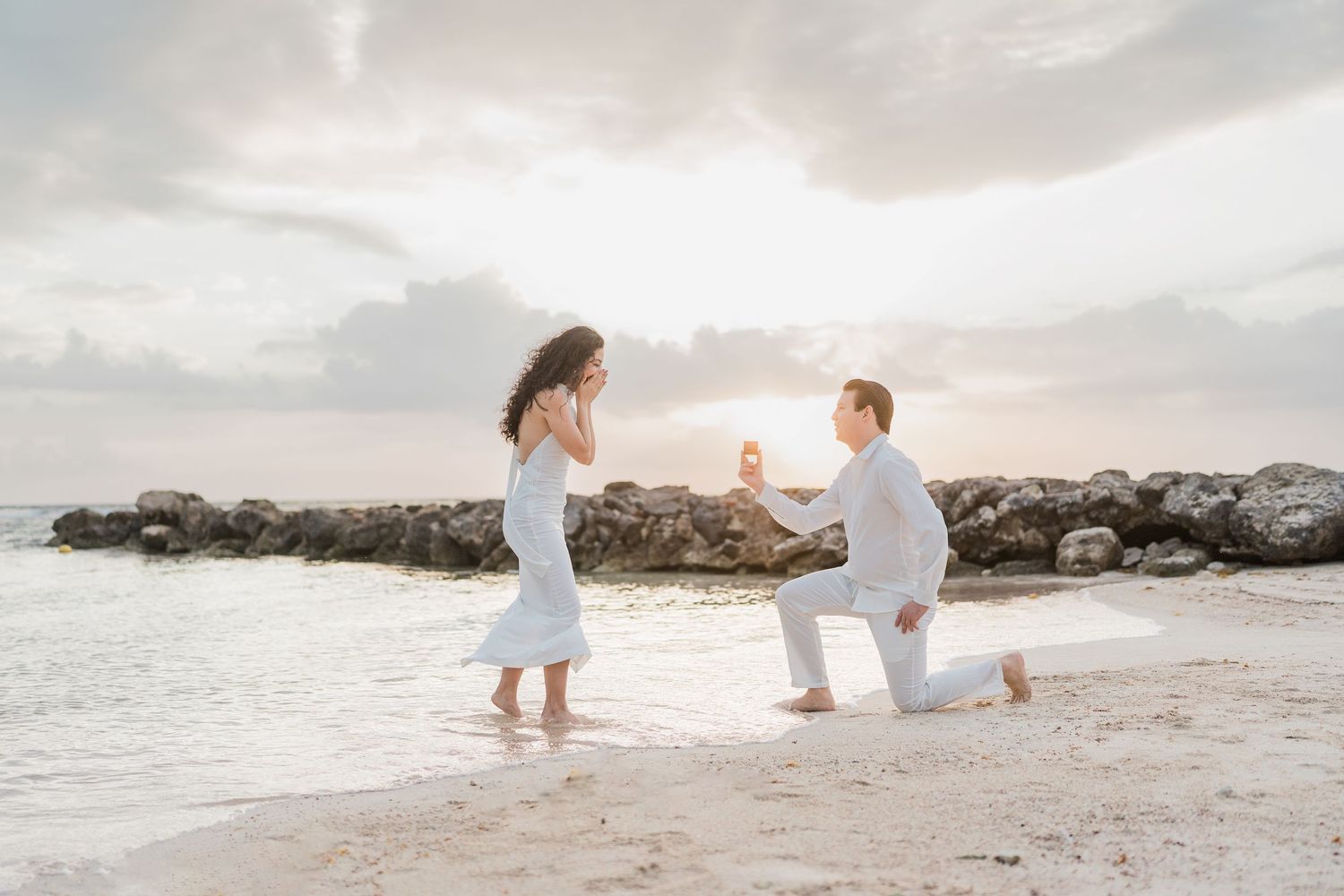 Man proposes to woman on a sandy beach at sunset, both dressed in white, with rocky shoreline in the background.