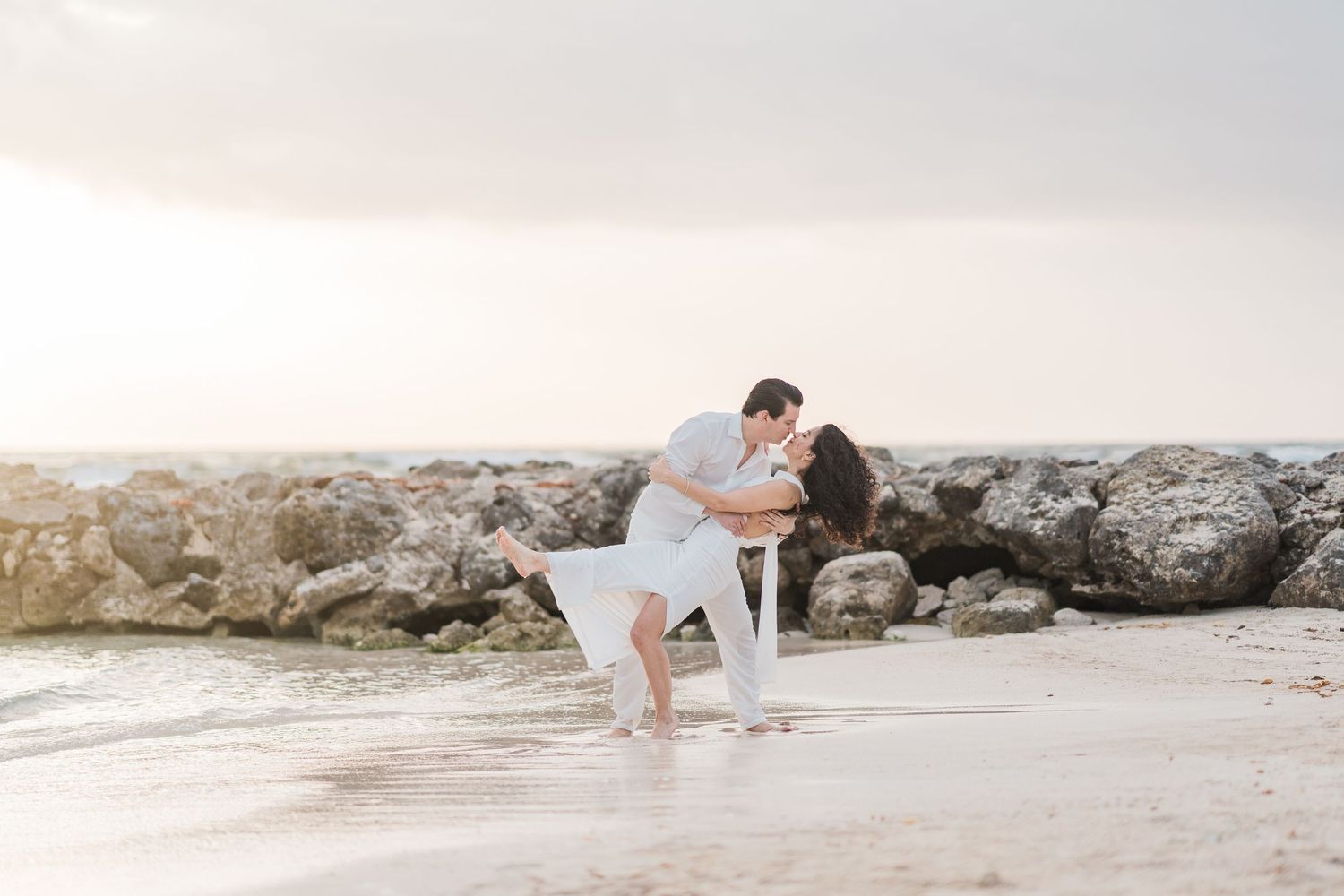 Couple sharing a romantic dip kiss on a sandy beach with rocky shoreline and soft pastel sky at sunset.