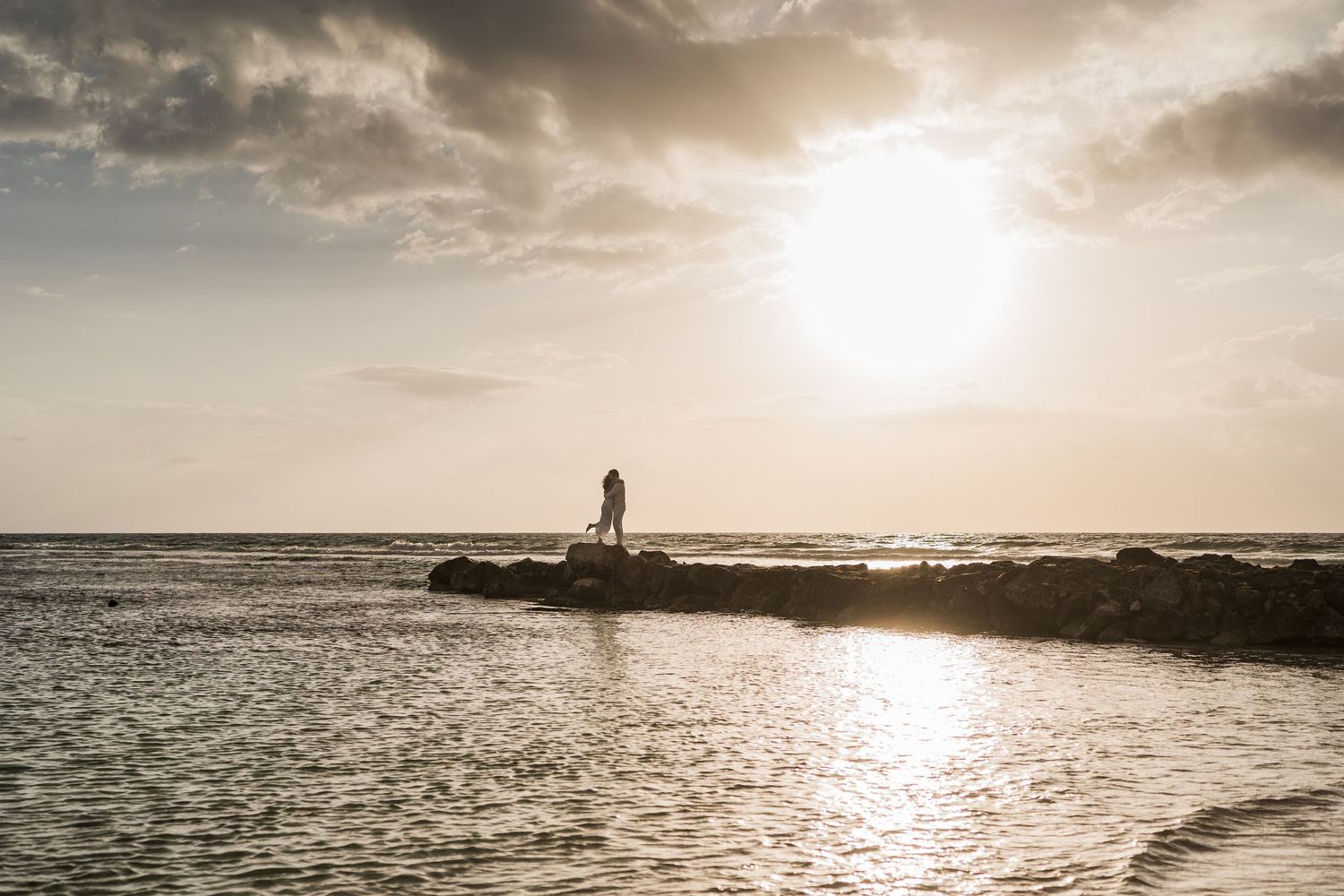 Silhouette of a person standing on rocky jetty at sunset over calm ocean waters with golden sky.