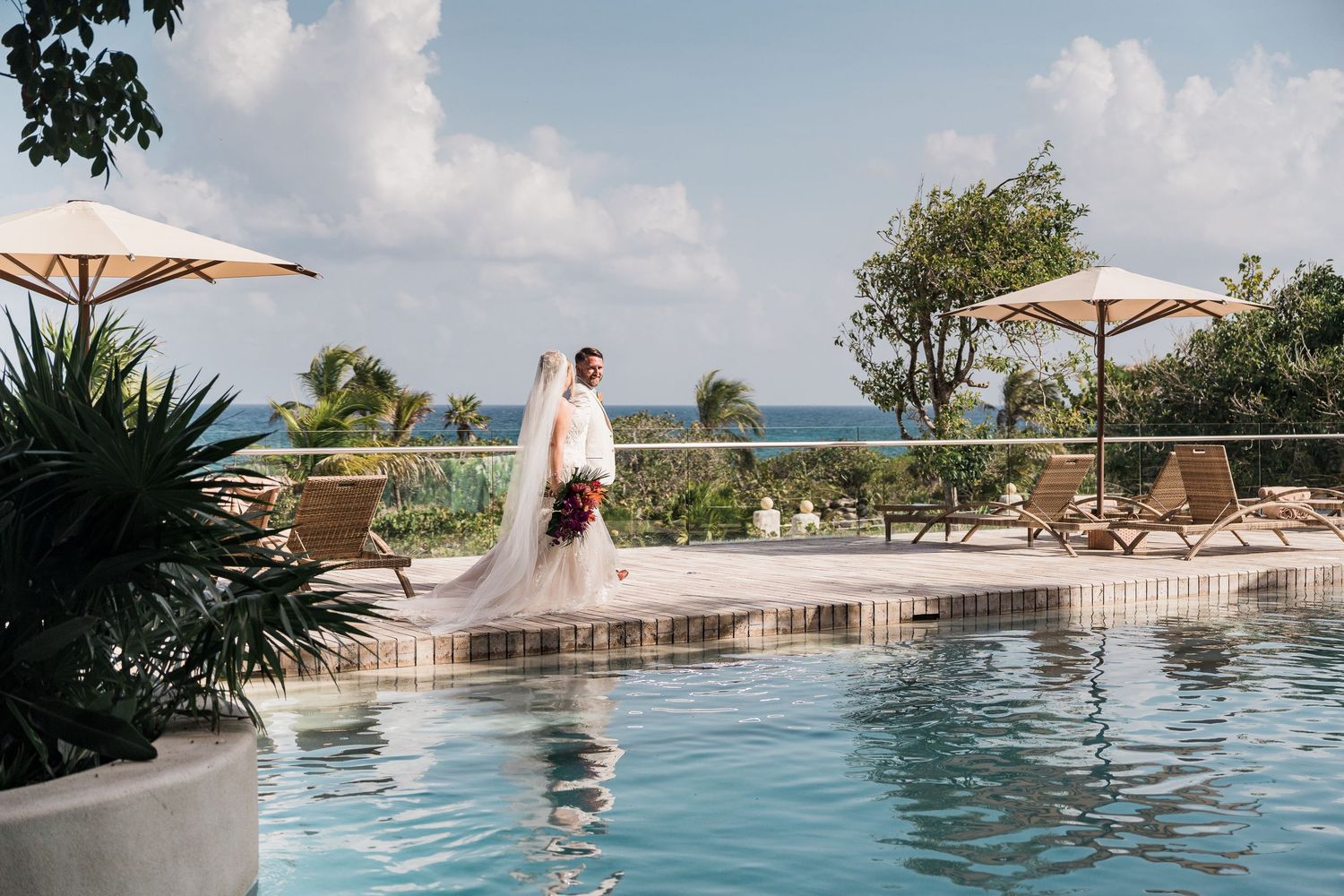 Hotel Xcaret México Wedding  - Bride and groom pose by a resort pool with ocean views and tropical landscaping on their wedding day.