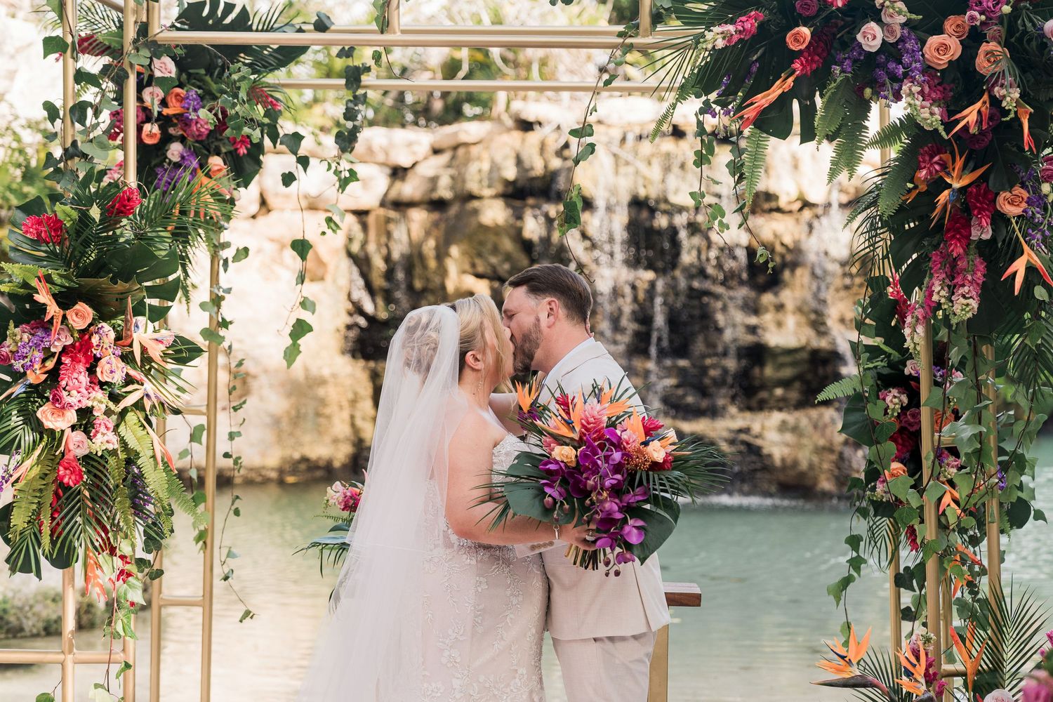 Bride and groom kissing under a vibrant floral arch with tropical flowers and waterfall backdrop at outdoor wedding.