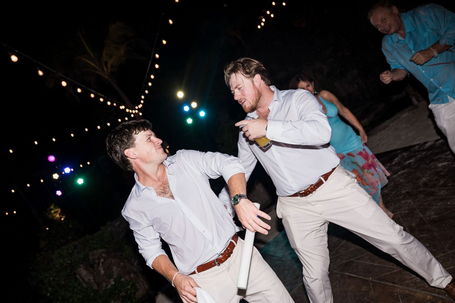Two men in white shirts dancing energetically at an outdoor evening wedding reception with string lights.