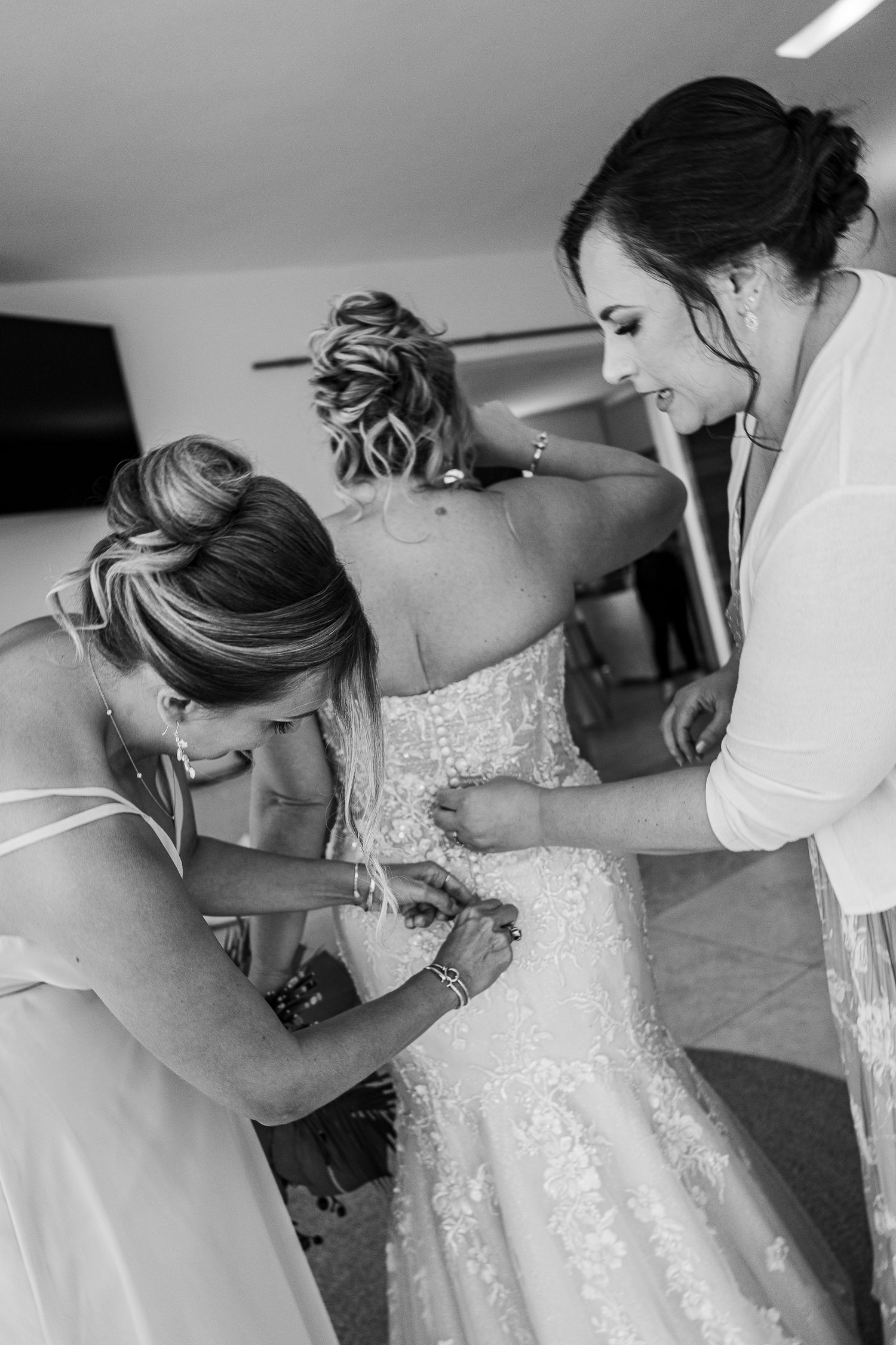 Hotel Xcaret México Wedding  - Two bridesmaids help a bride fasten her lace wedding dress in a black and white bridal preparation photo.
