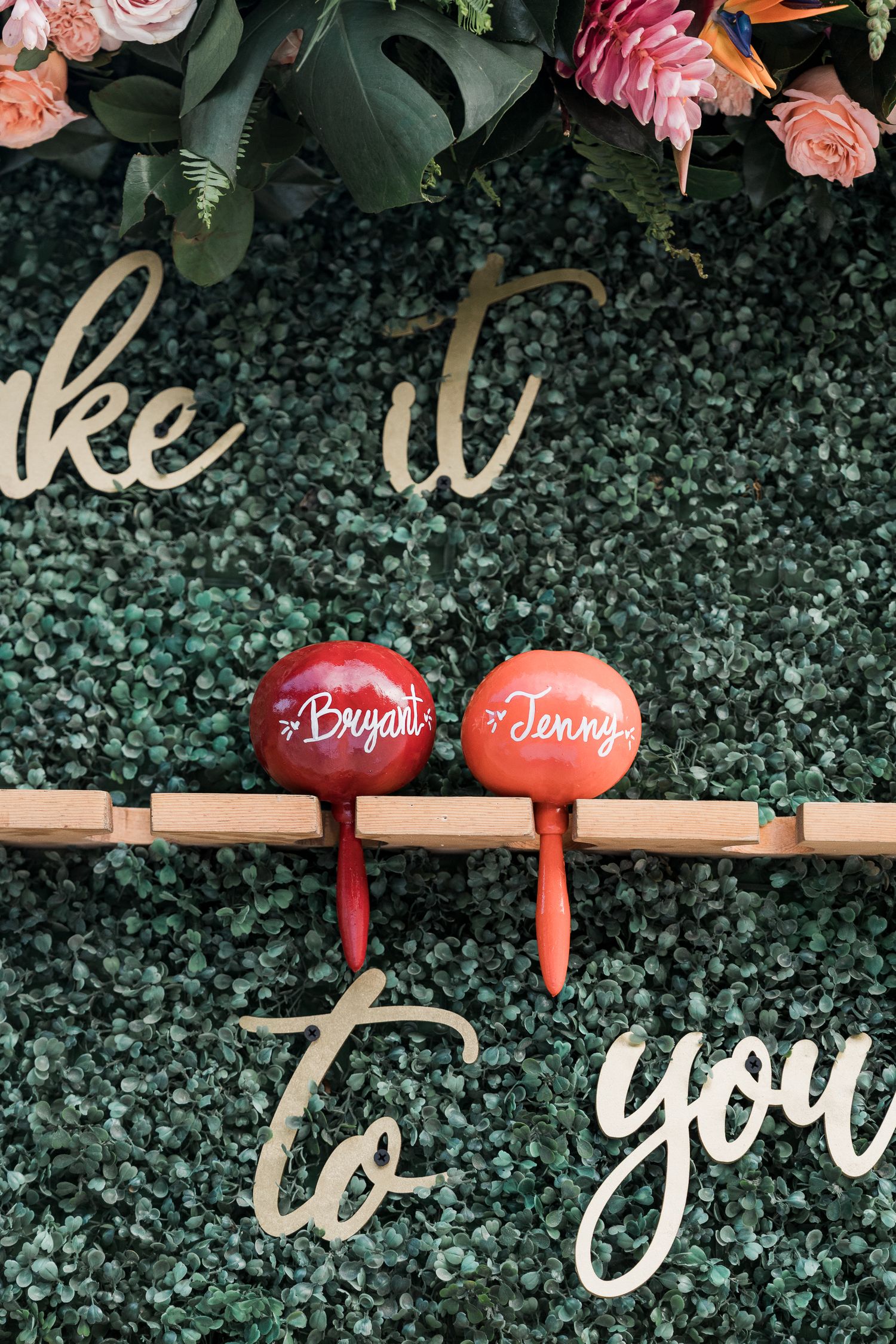 Personalized red maracas for Bryan and Jenny displayed on a greenery wall with floral decor and gold script lettering.