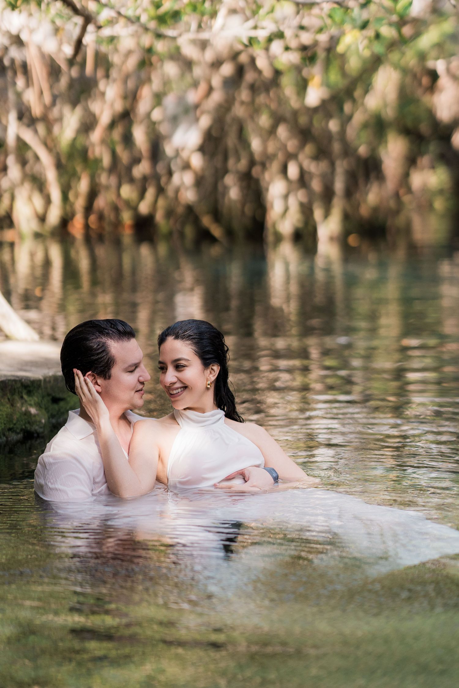 A couple in white wedding attire embraces romantically while wading in a serene, tree-lined natural pool.