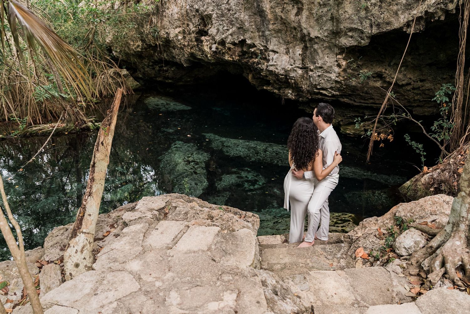 Couple embracing at the entrance of a lush cenote cave with rocky steps and tropical greenery in Mexico.