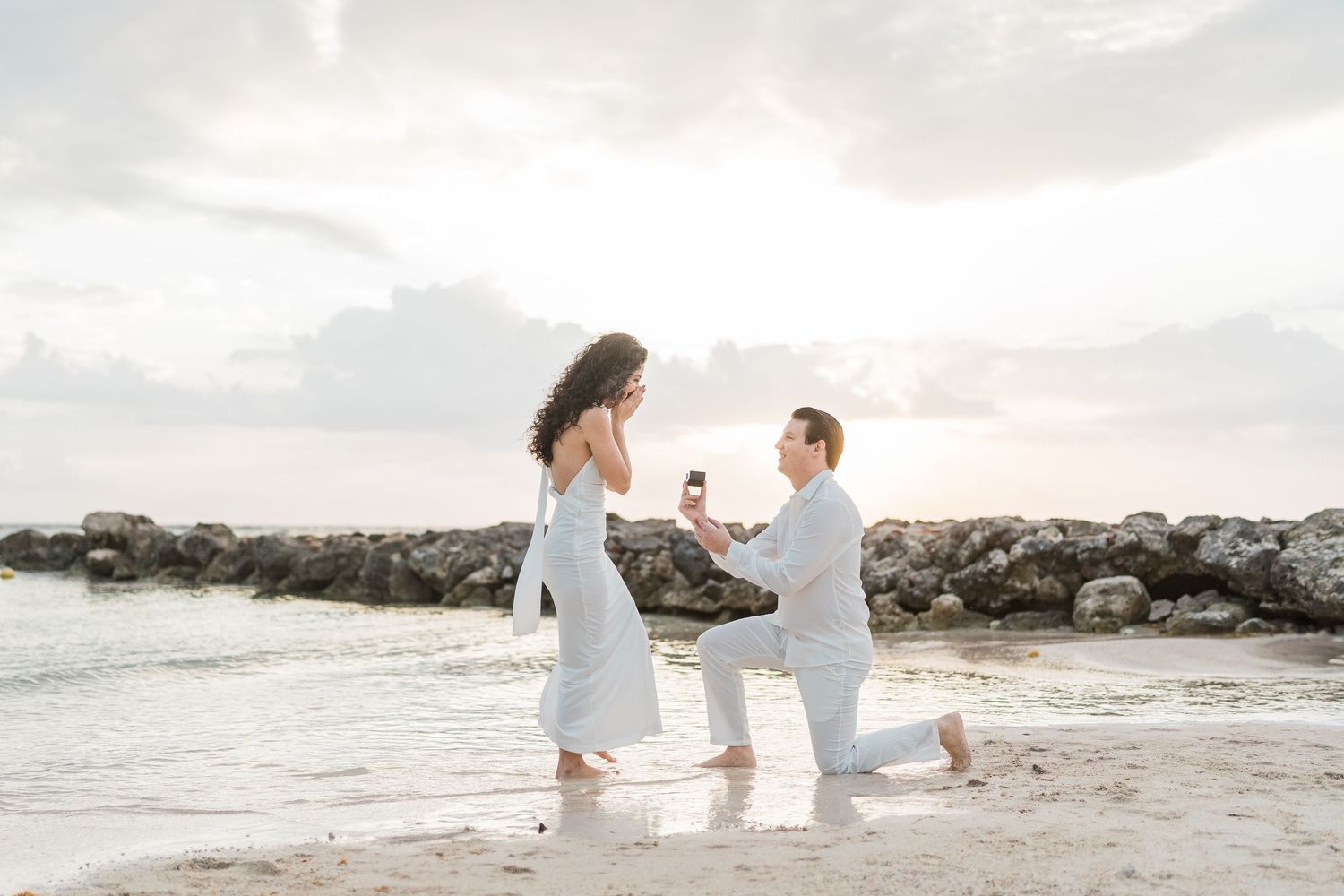 Man proposes to woman on a sandy beach at sunset, kneeling with a ring while waves lap the shore.