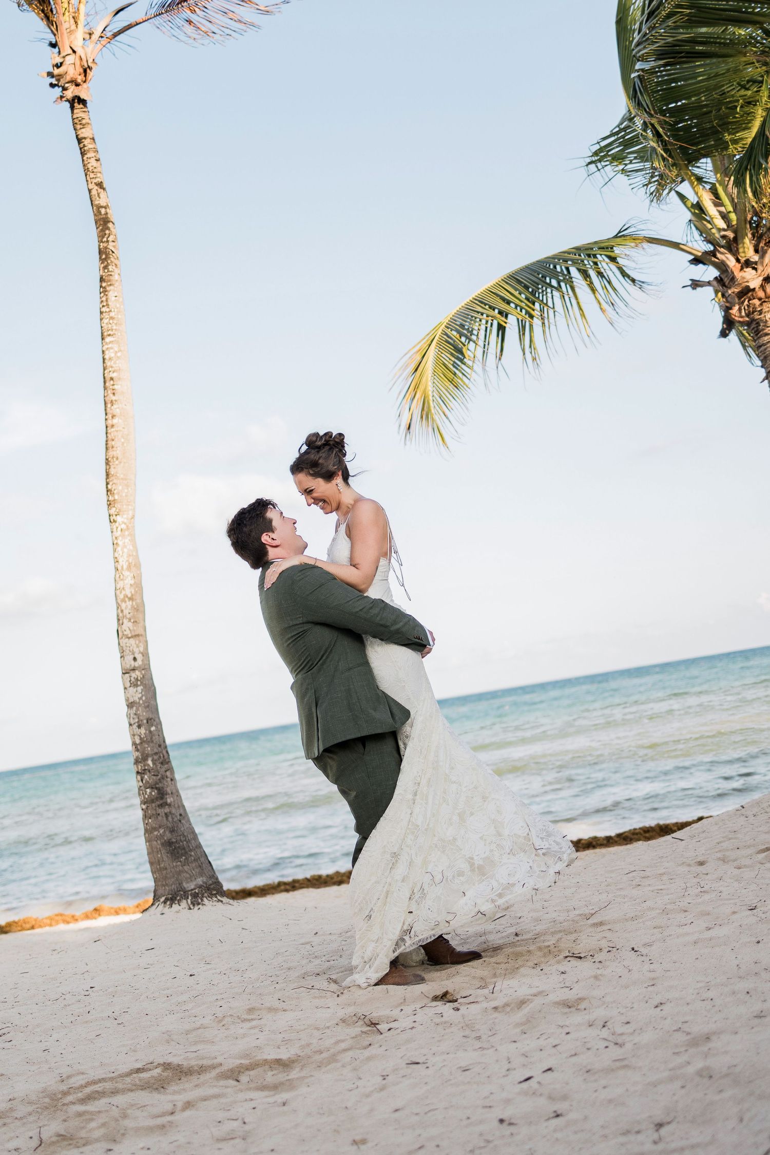 Groom lifting bride in white lace dress on tropical beach with palm trees and turquoise ocean backdrop.