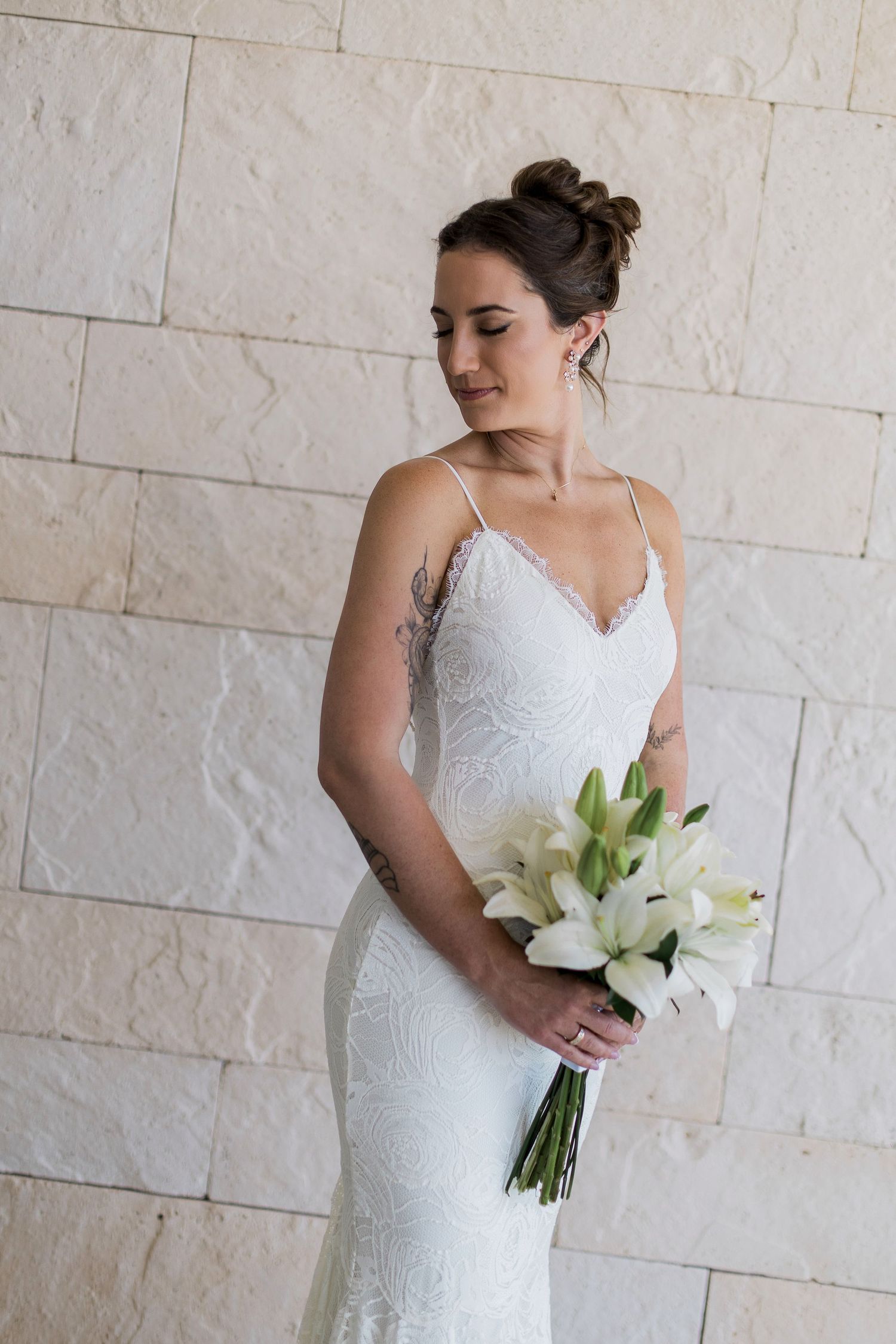 Bride in white lace spaghetti strap wedding gown holding white lily bouquet, standing against stone wall.