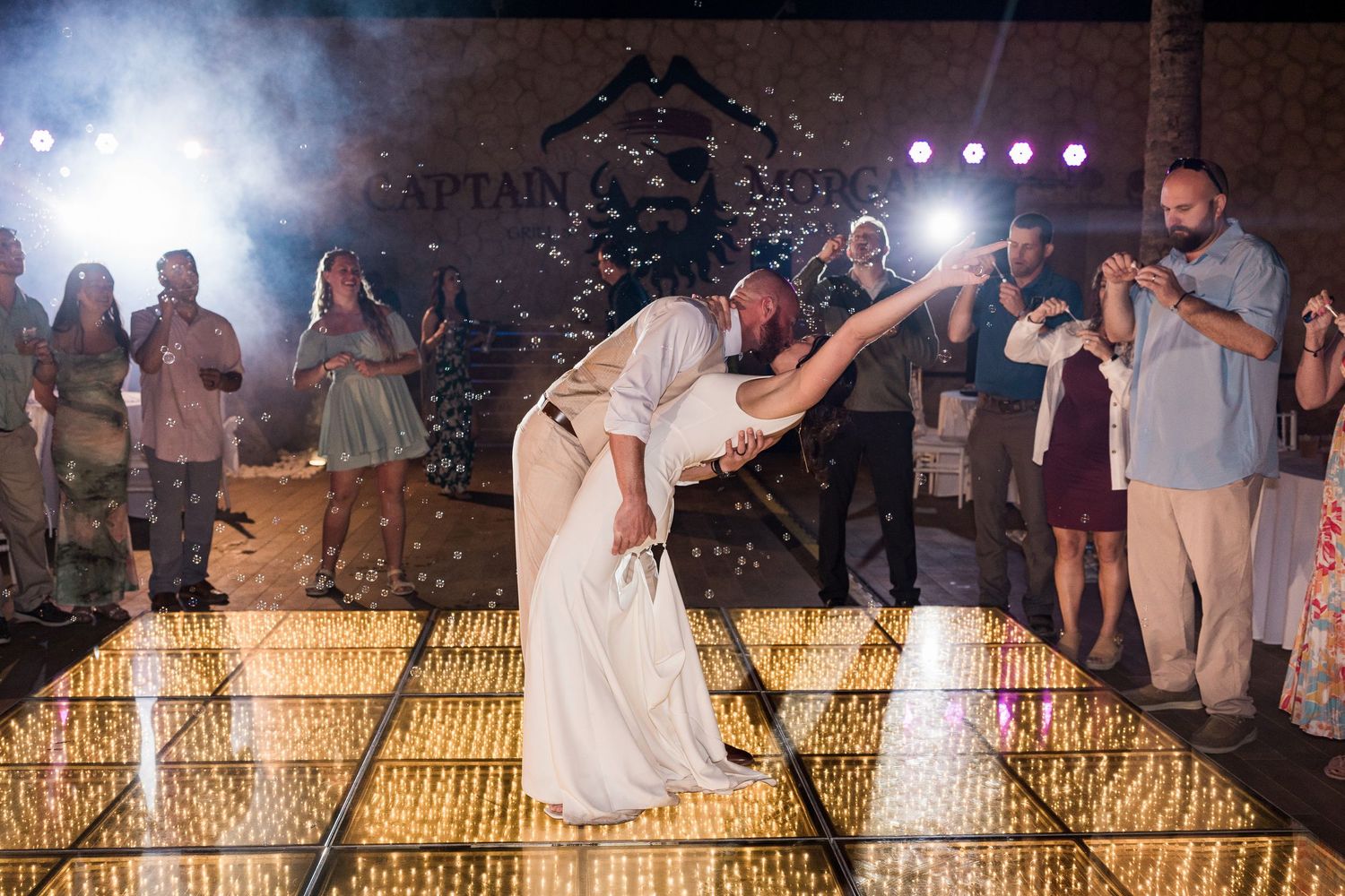 Bride and groom share romantic dip dance on illuminated floor at outdoor wedding reception with guests watching.
