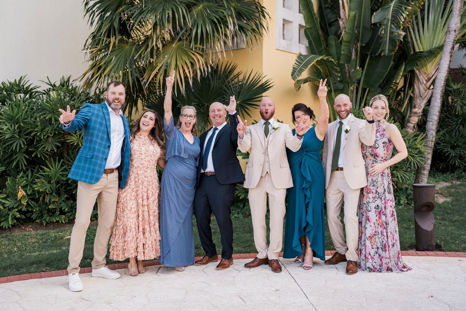 Group of eight friends posing joyfully outdoors near tropical palm trees at a wedding celebration.