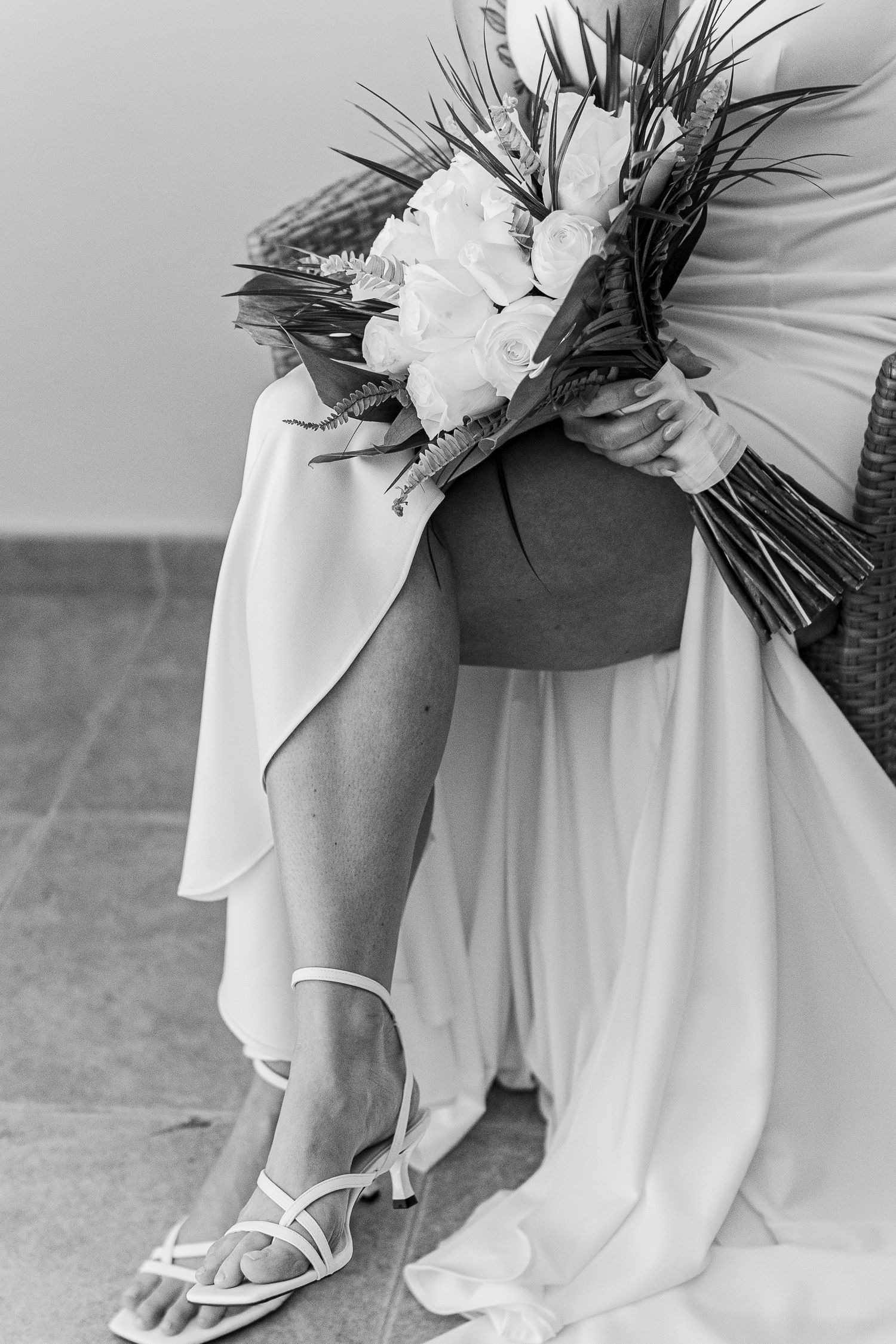 Black and white photo of a bride holding a floral bouquet, wearing a white dress and strappy heels while seated.