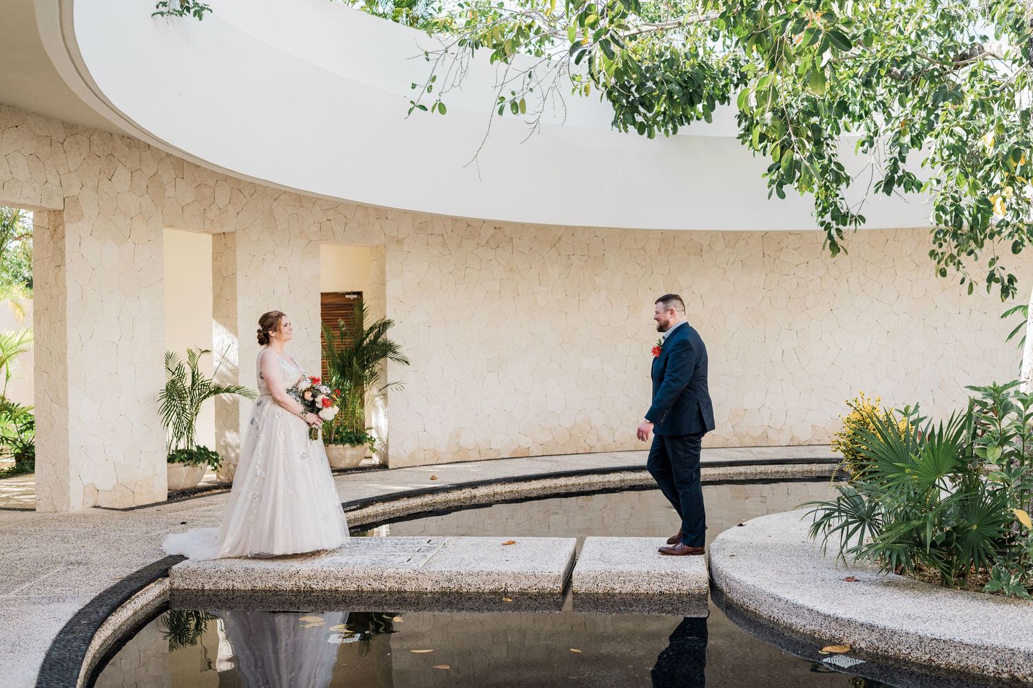 Secrets Akumal Wedding - Bride and groom share a first look moment by a reflective pool in an elegant outdoor wedding venue.