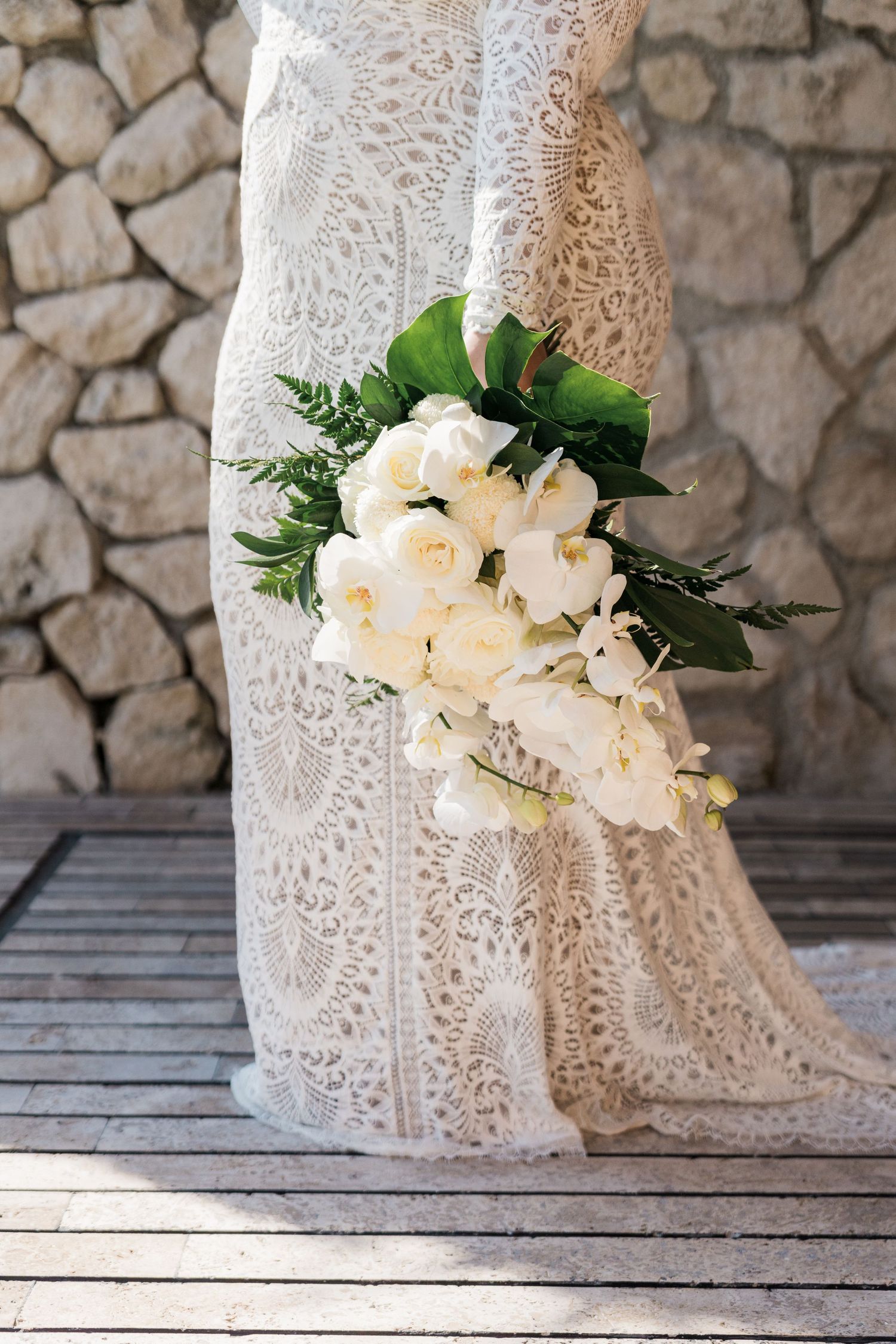 Xcaret Arte Wedding - Bride in lace wedding gown holding cascading white rose and orchid bouquet with green foliage against stone wall.