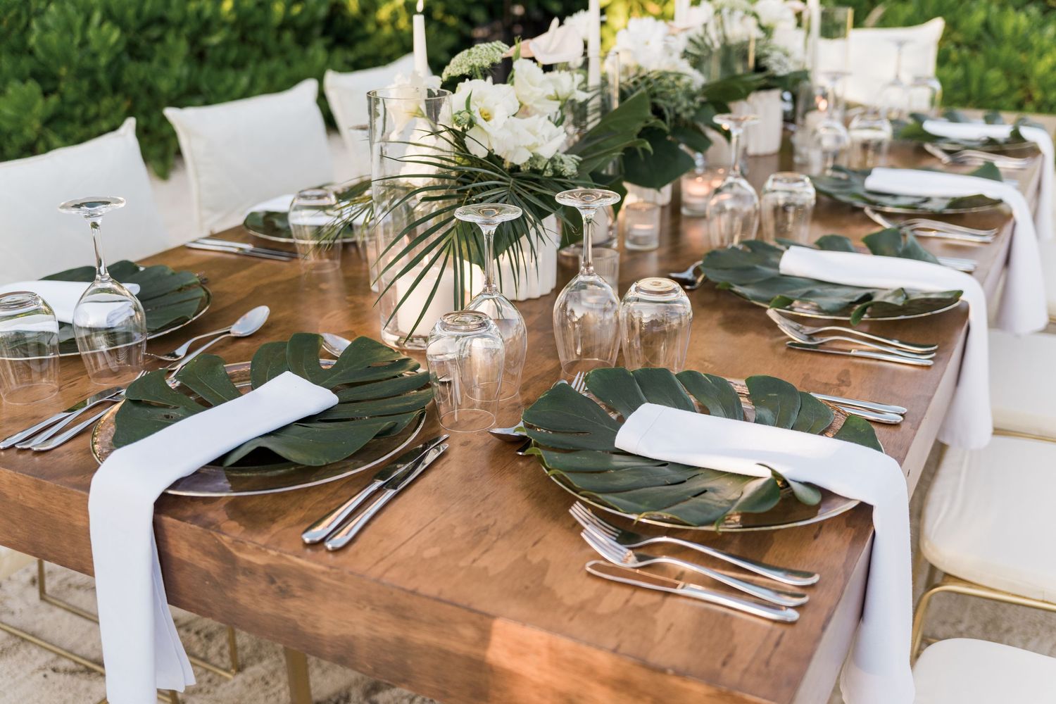 Elegant outdoor wedding table setting with monstera leaf chargers, white napkins, candles, and floral centerpieces on rustic wood table.