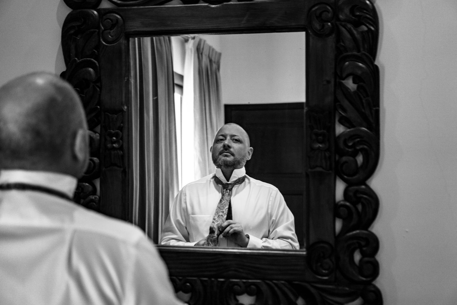 Black and white photo of bald groom adjusting tie while looking in ornate mirror.