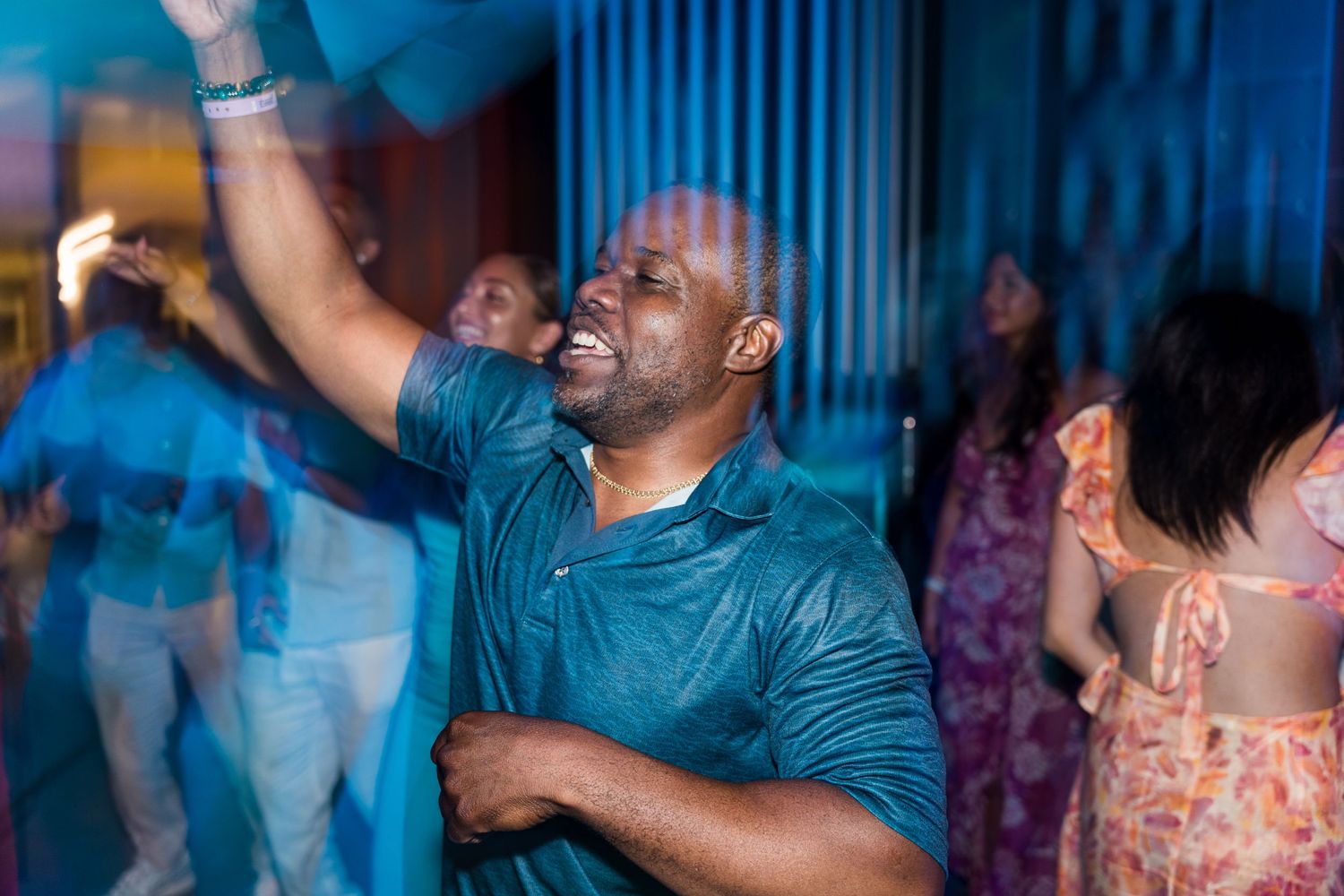 Man in teal shirt dancing enthusiastically with raised arm at a wedding party.