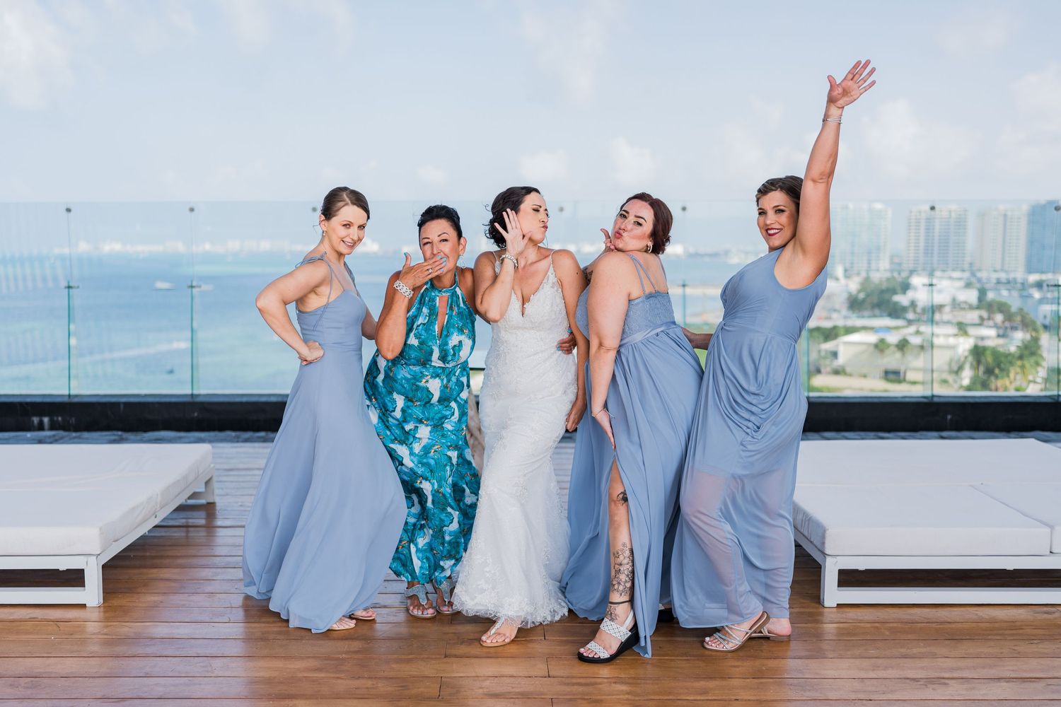 Bride and bridesmaids in blue dresses celebrating on rooftop with ocean views.
