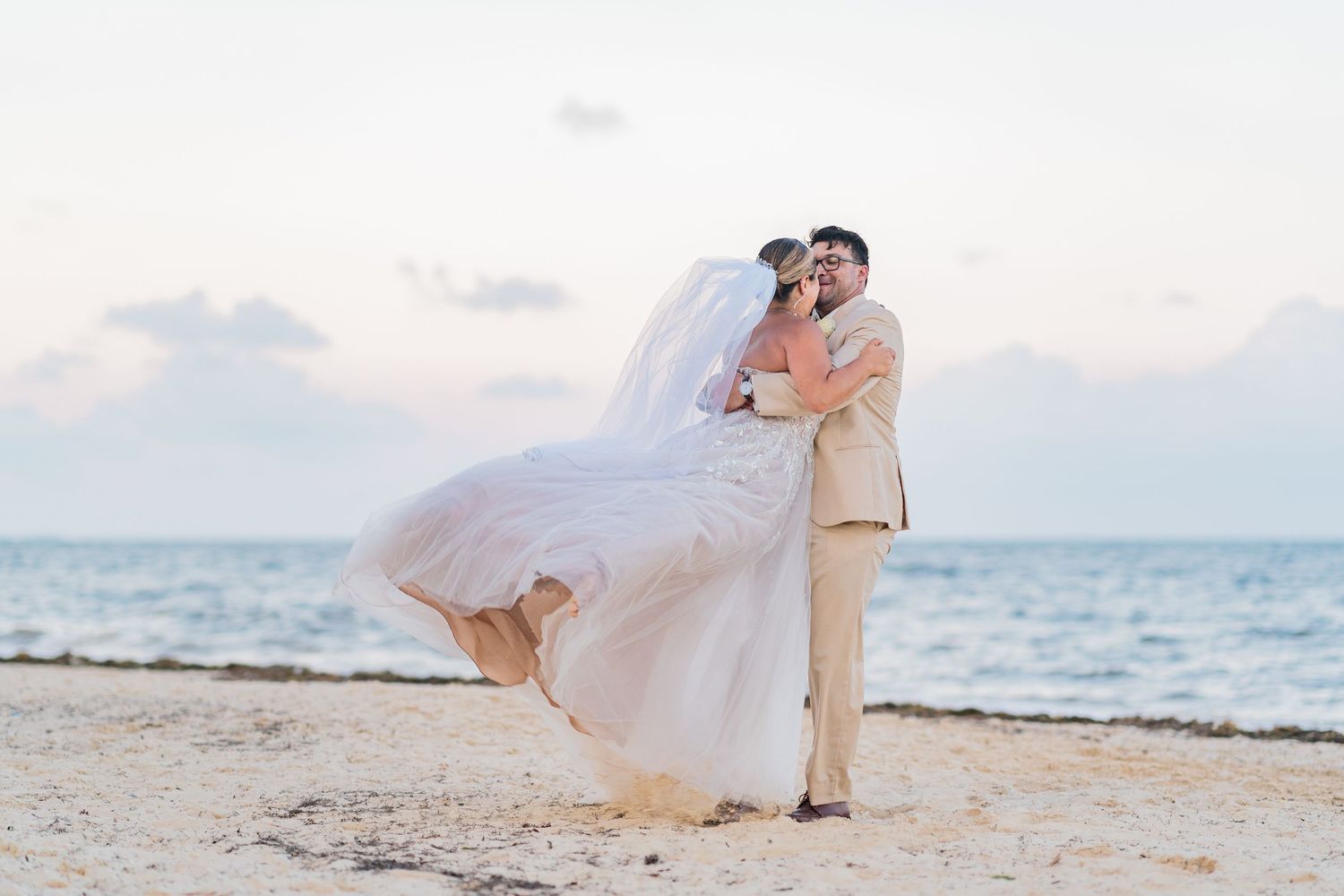 Groom lifting bride in flowing white gown on a serene beach at sunset.