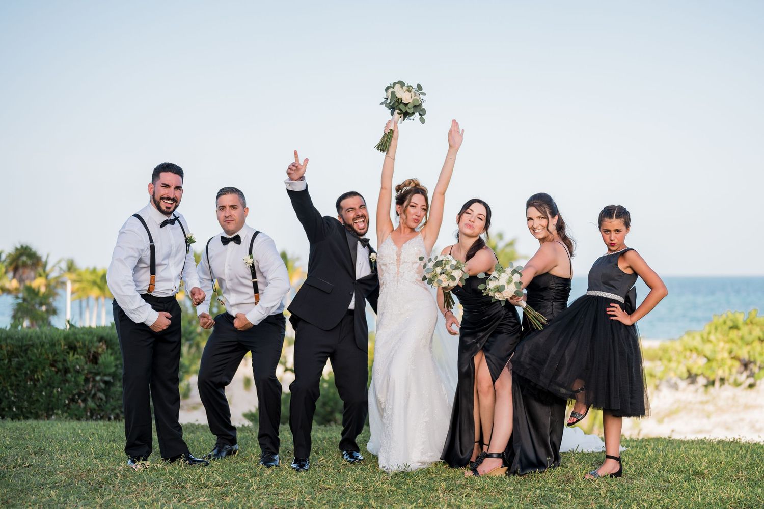 Wedding party celebrates outdoors near the beach, bridesmaids in black dresses and groomsmen in suspenders.