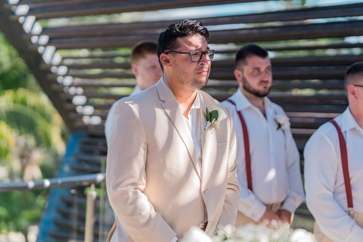 Groom in beige suit with glasses waits emotionally at outdoor wedding ceremony altar.