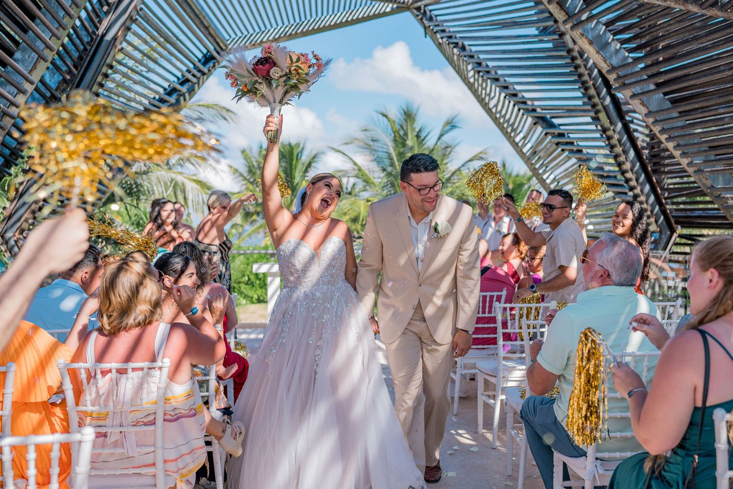 Newlyweds walk down the aisle as guests cheer under a rustic wooden arch at a tropical beach wedding.