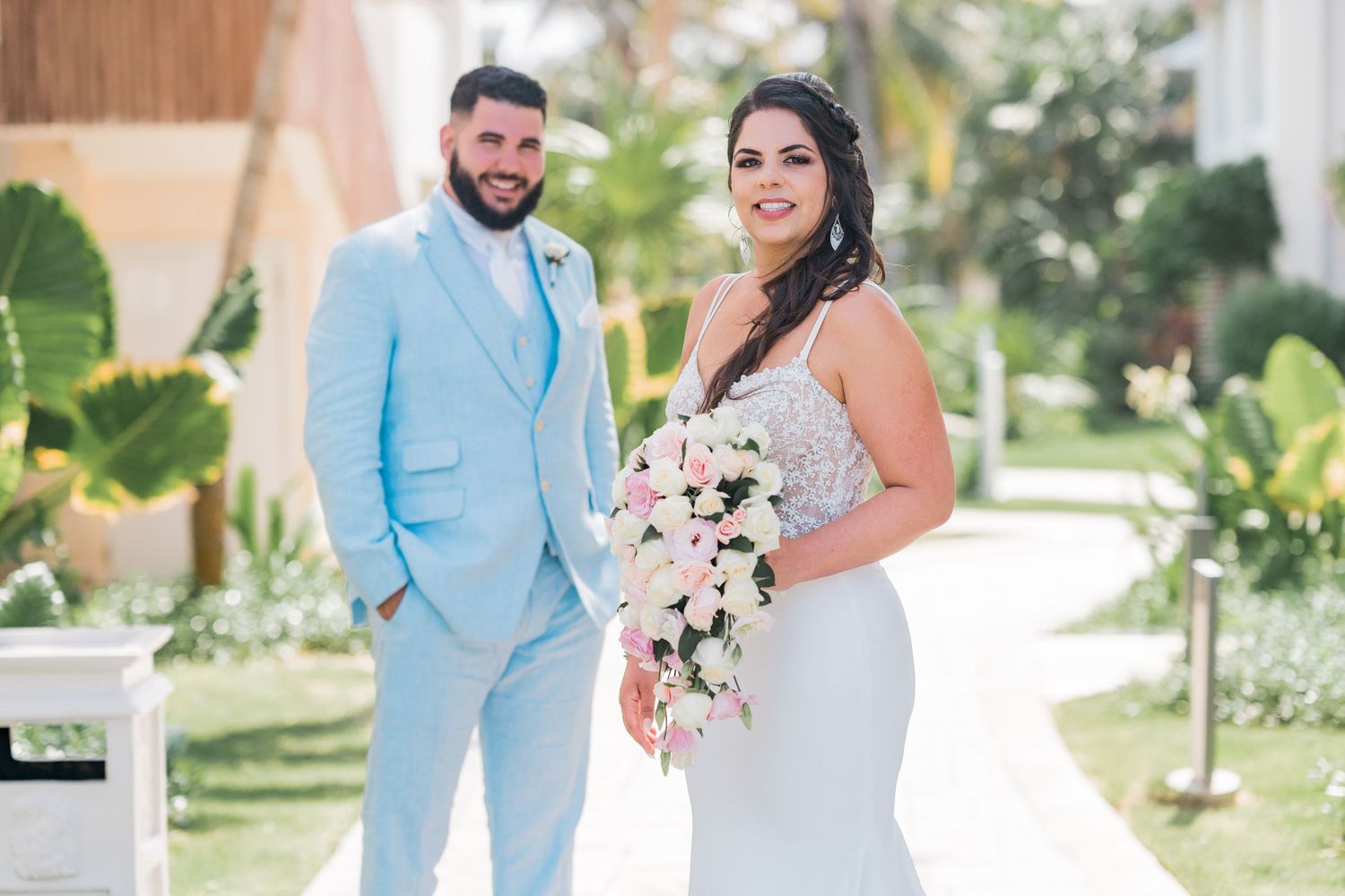 Bride and groom posing together outdoors at a tropical resort, bride holding a cascading floral bouquet.