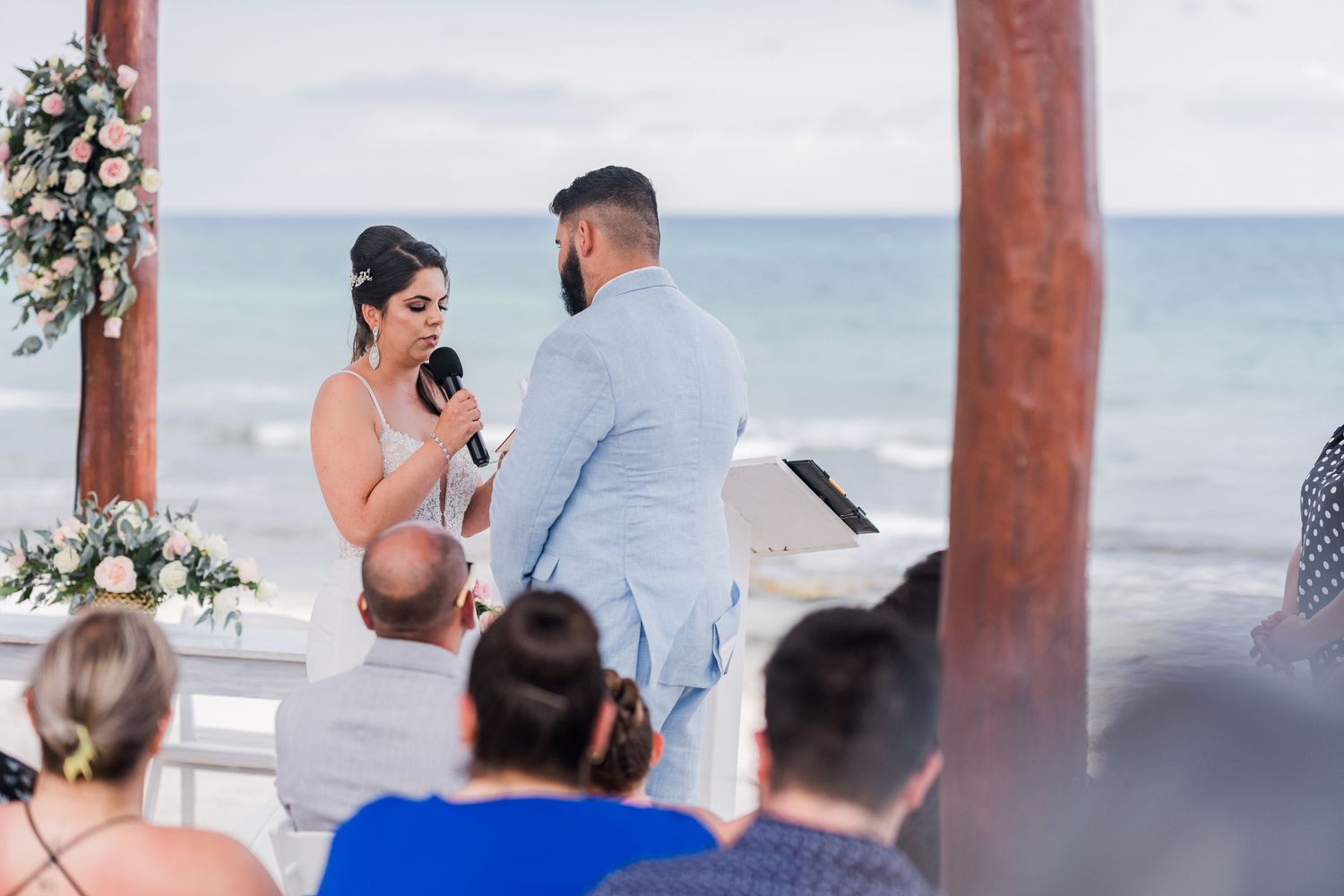 Bride reads vows into microphone facing groom at romantic outdoor beach wedding with ocean backdrop.