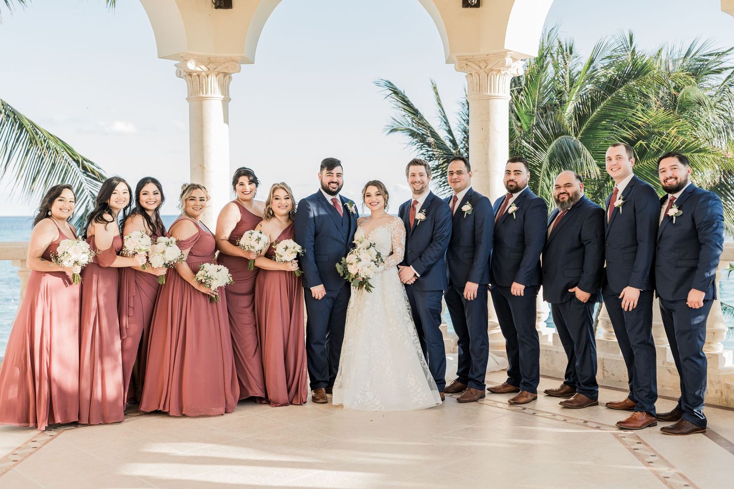 Full wedding party with bridesmaids in rose gowns and groomsmen in navy suits posing at beachside venue.