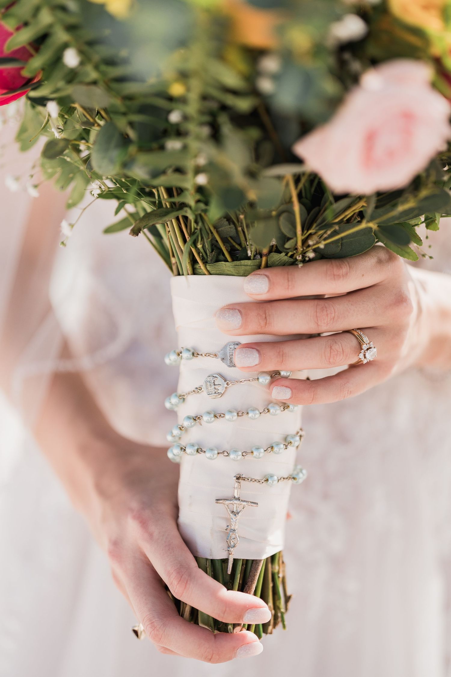 Bride holds floral bouquet wrapped in white ribbon with pearl rosary and diamond engagement ring.