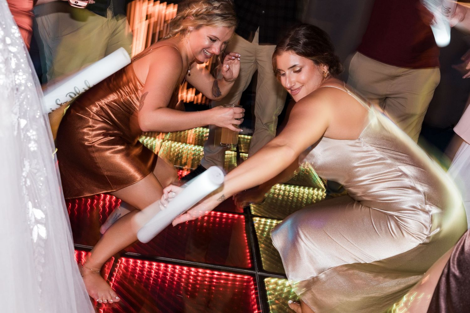 Two bridesmaids laugh and dance together on a glowing LED dance floor at a wedding reception.