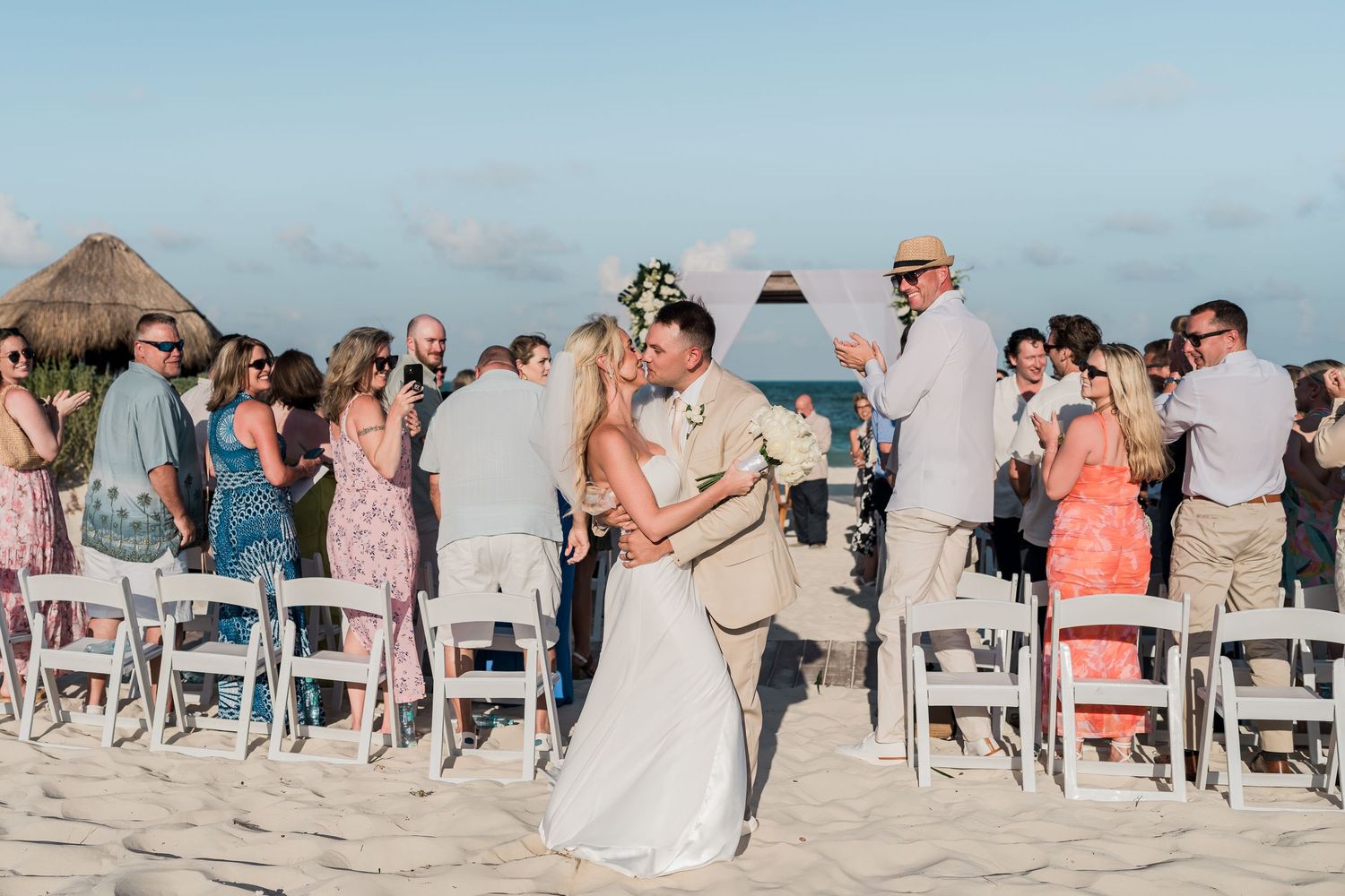 Newlywed couple shares romantic embrace on sandy beach as wedding guests celebrate around them at sunset.